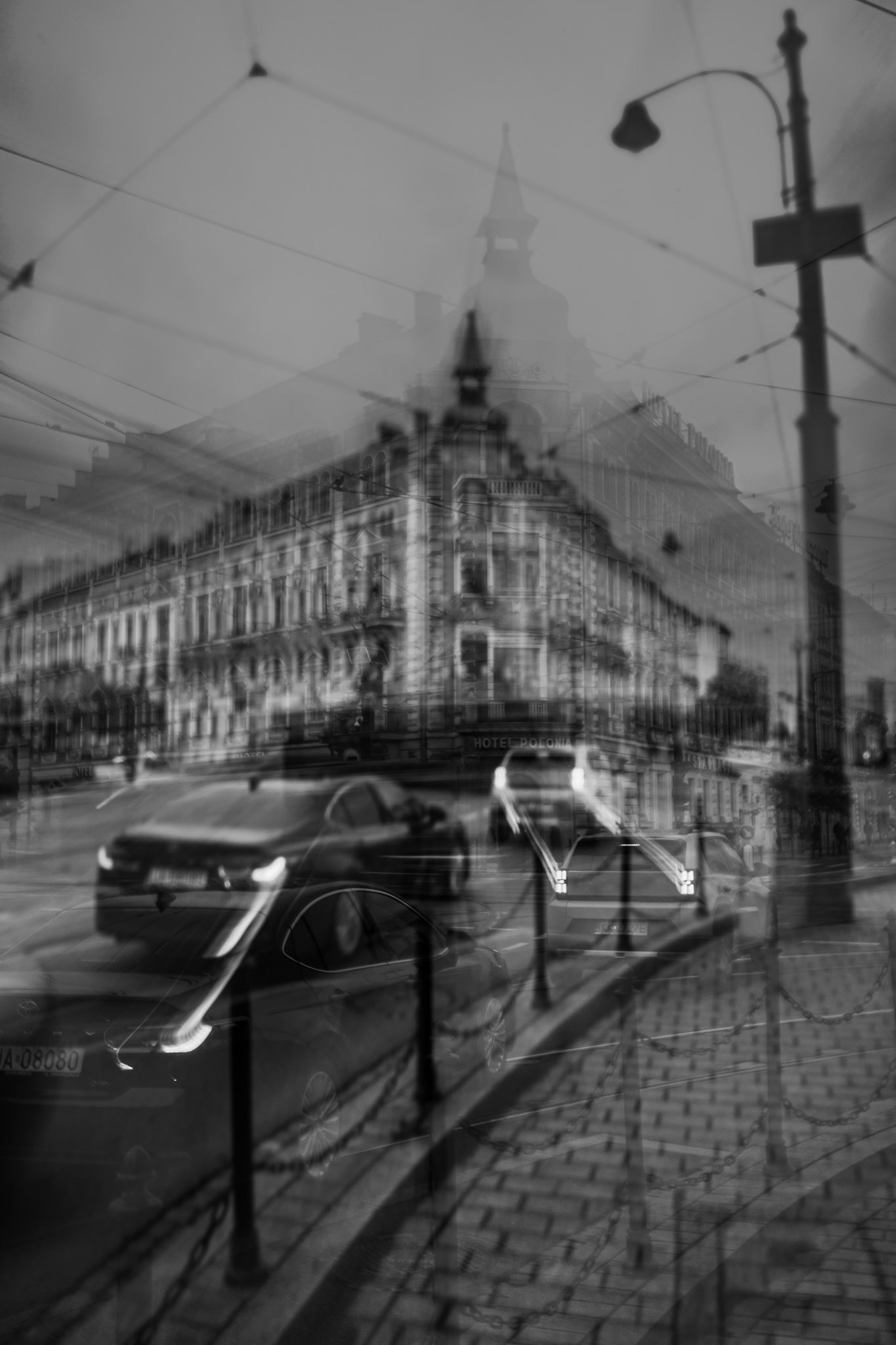 Moody black & white multiple-exposure of a city street, layering cars, tram wires, and the grand, historic Hotel Polonia into a surreal landscape.