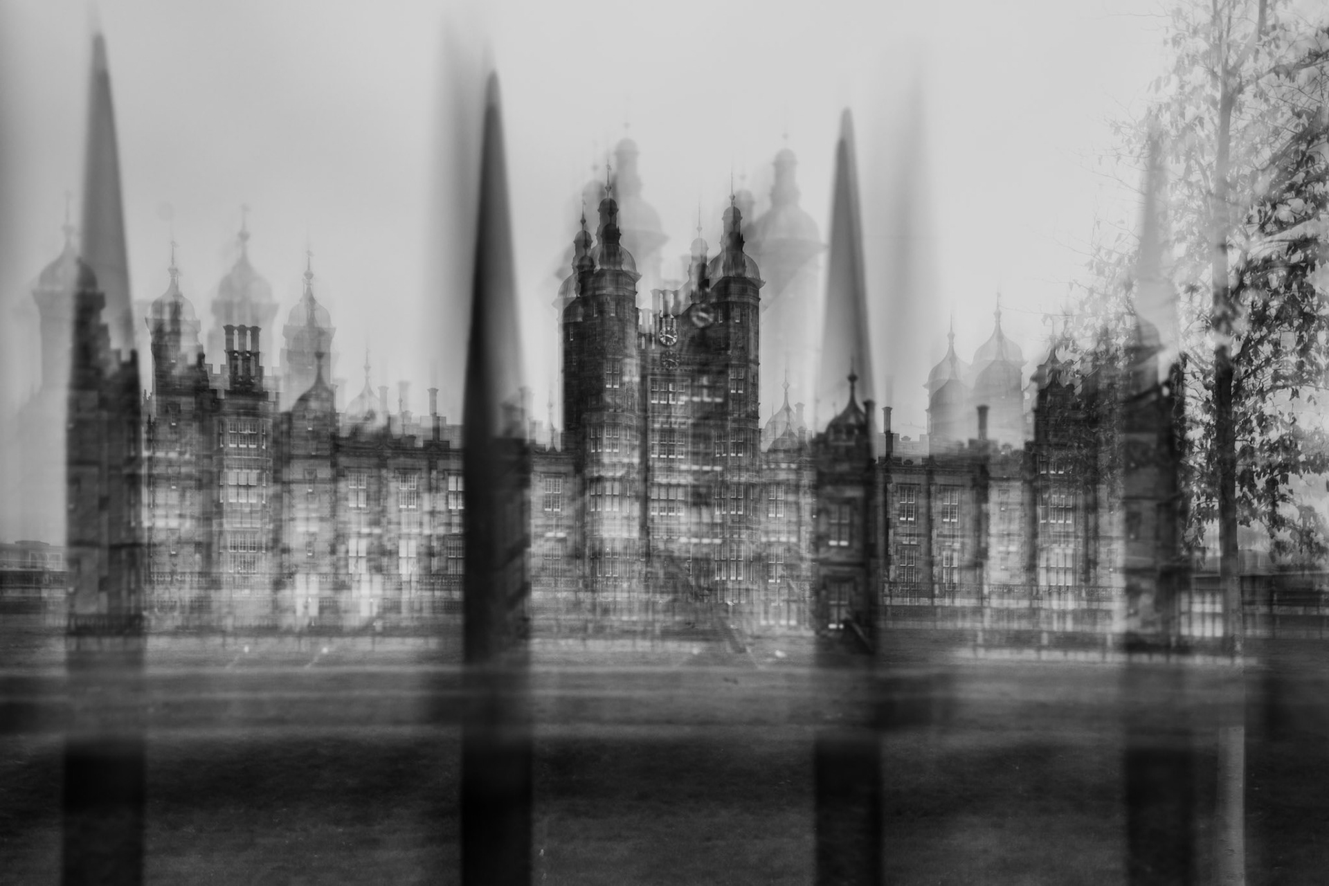 Black-and-white long exposure photograph of a historic Gothic-style building with ornate towers and domes, seen blurred behind tall iron fence spikes in the foreground.