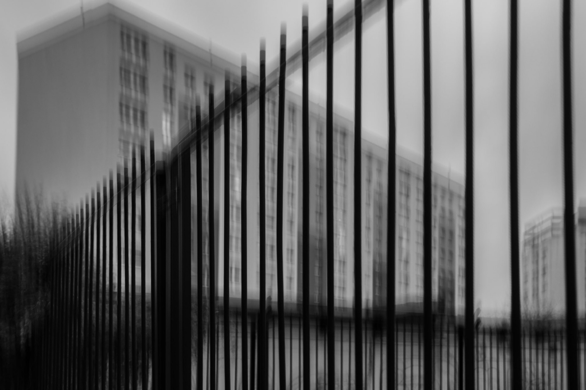 Abstract black and white ICM photography of a modern urban building seen through tall metal fence bars, creating a blurred motion effect and dramatic perspective.