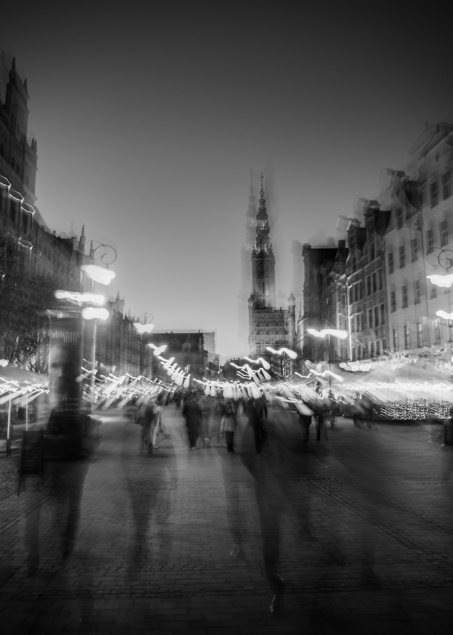 Blurred long-exposure view of a busy European city square at night with pedestrians and glowing street lights leading toward a tall historic tower