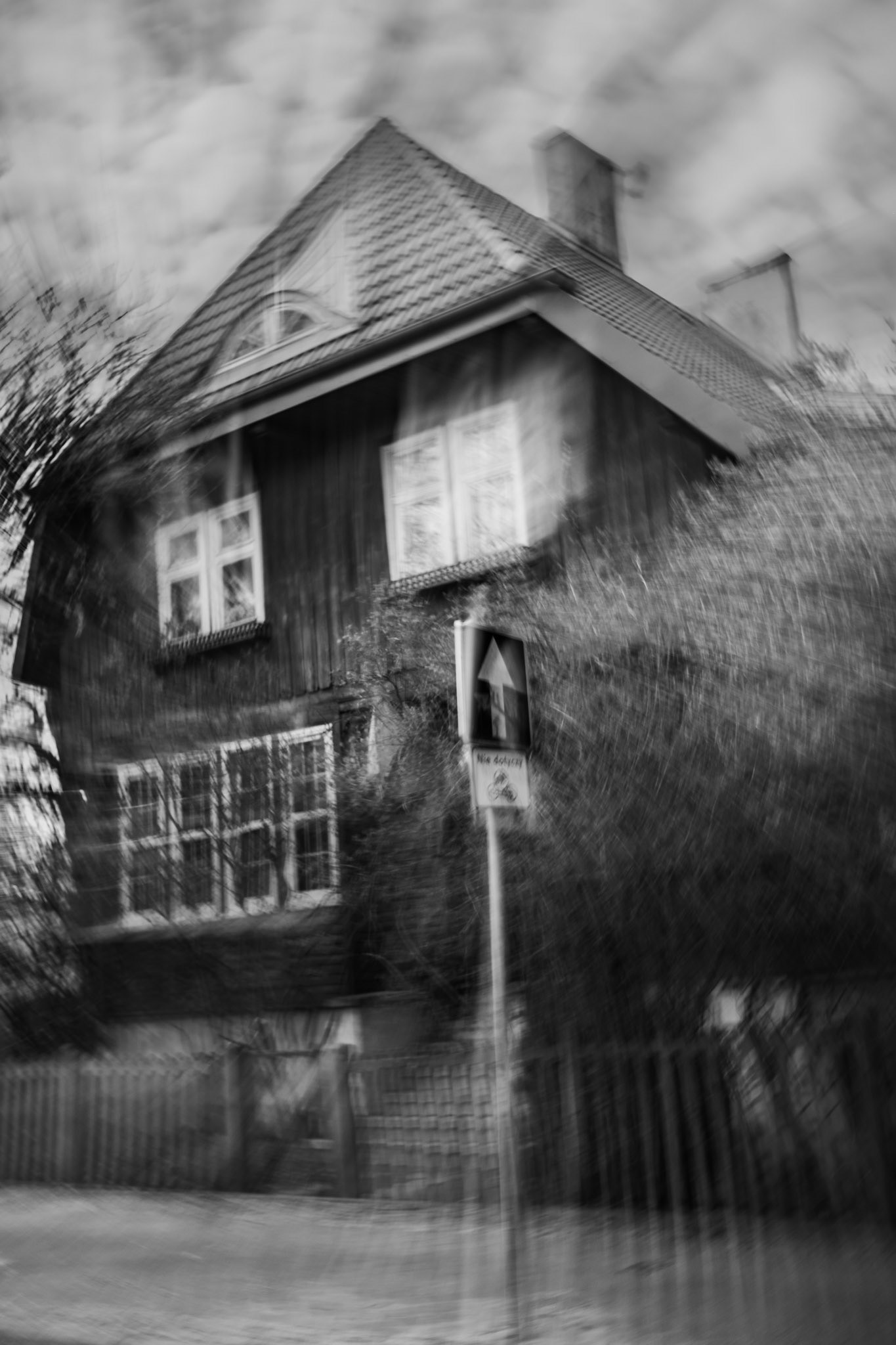 Blurry black-and-white photo of an old wooden house with a steep tiled roof, surrounded by trees and a fence, with a street sign in the foreground and a slightly distorted, eerie motion effect.