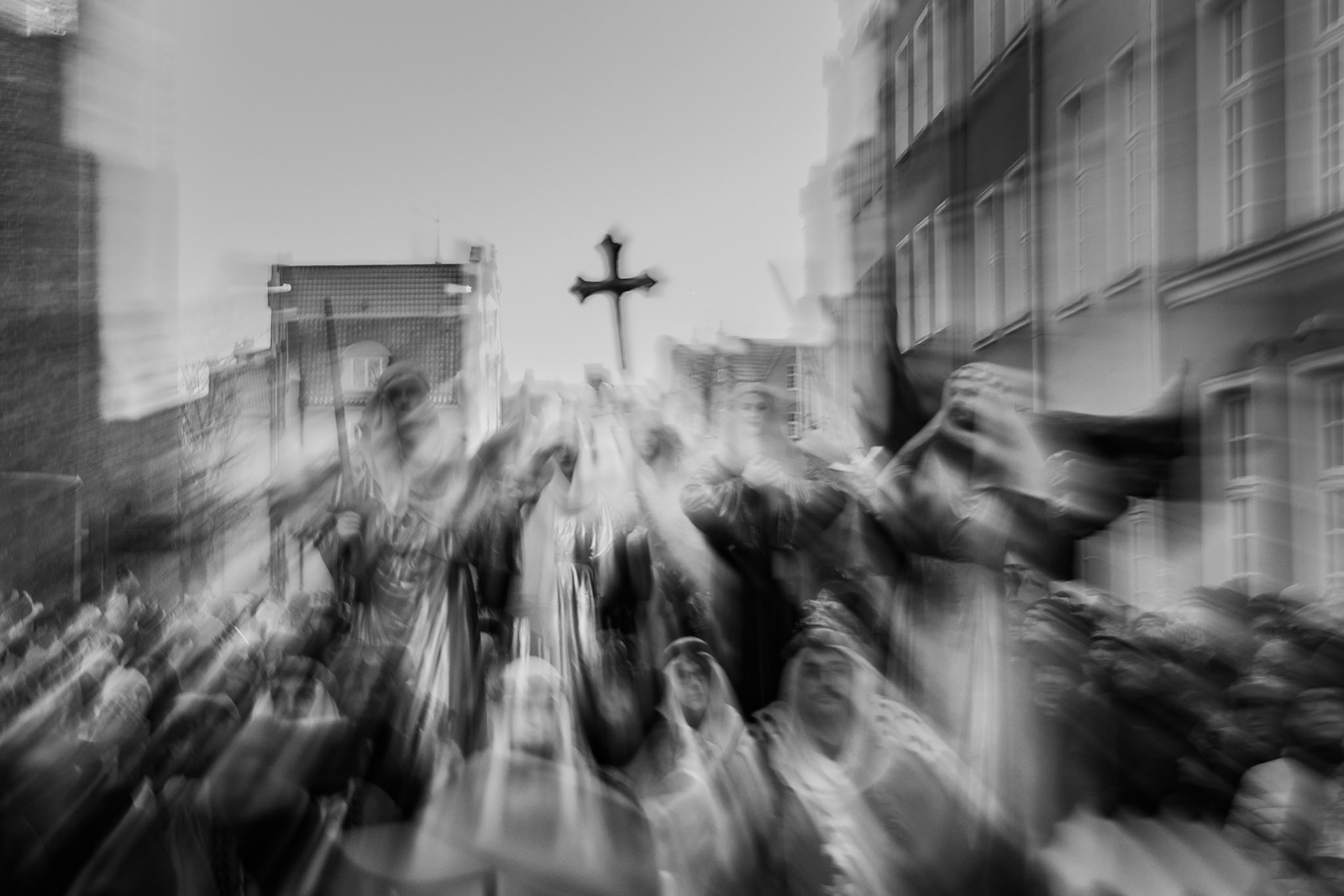 Motion-blurred black and white image of a religious procession, with worshippers carrying a cross through an urban street.