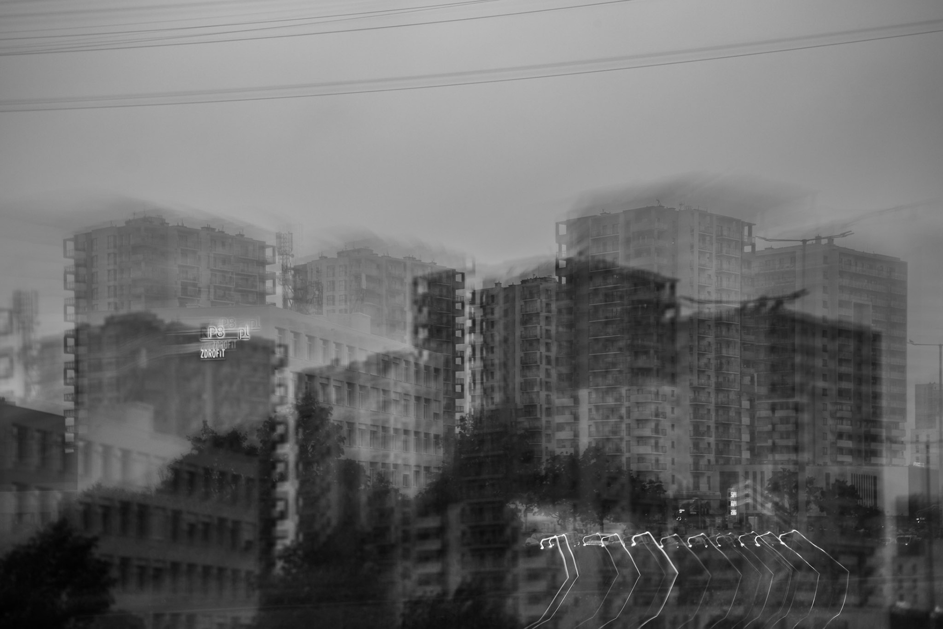 Black and white photo of a cityscape with tall apartment buildings, captured in ICM technique with a motion blur effect creating a ghostly, dreamlike appearance under a cloudy sky.