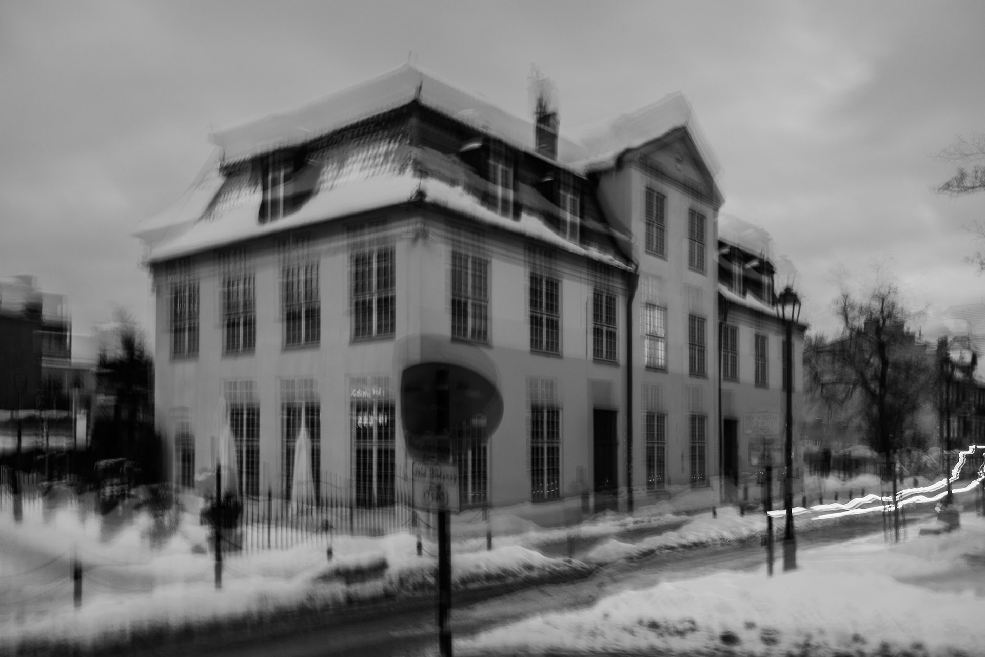Blurry black-and-white long-exposure photo of a historic European-style corner building in winter, with snow-covered streets and light trails from passing cars.