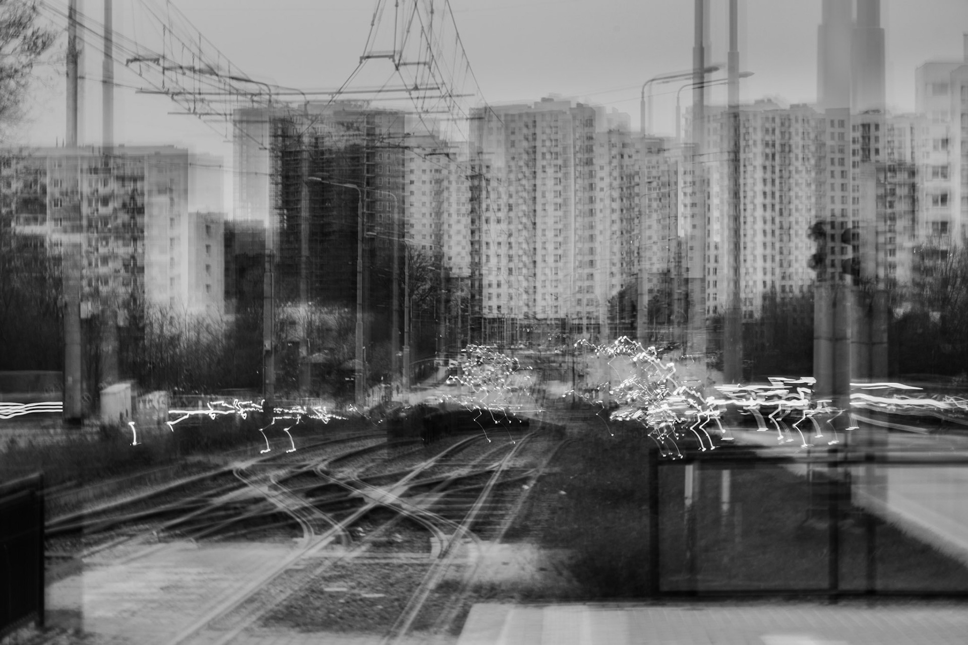 Blurred black-and-white cityscape with railway tracks, overhead power lines, and high-rise apartment buildings in the background, featuring motion light trails.