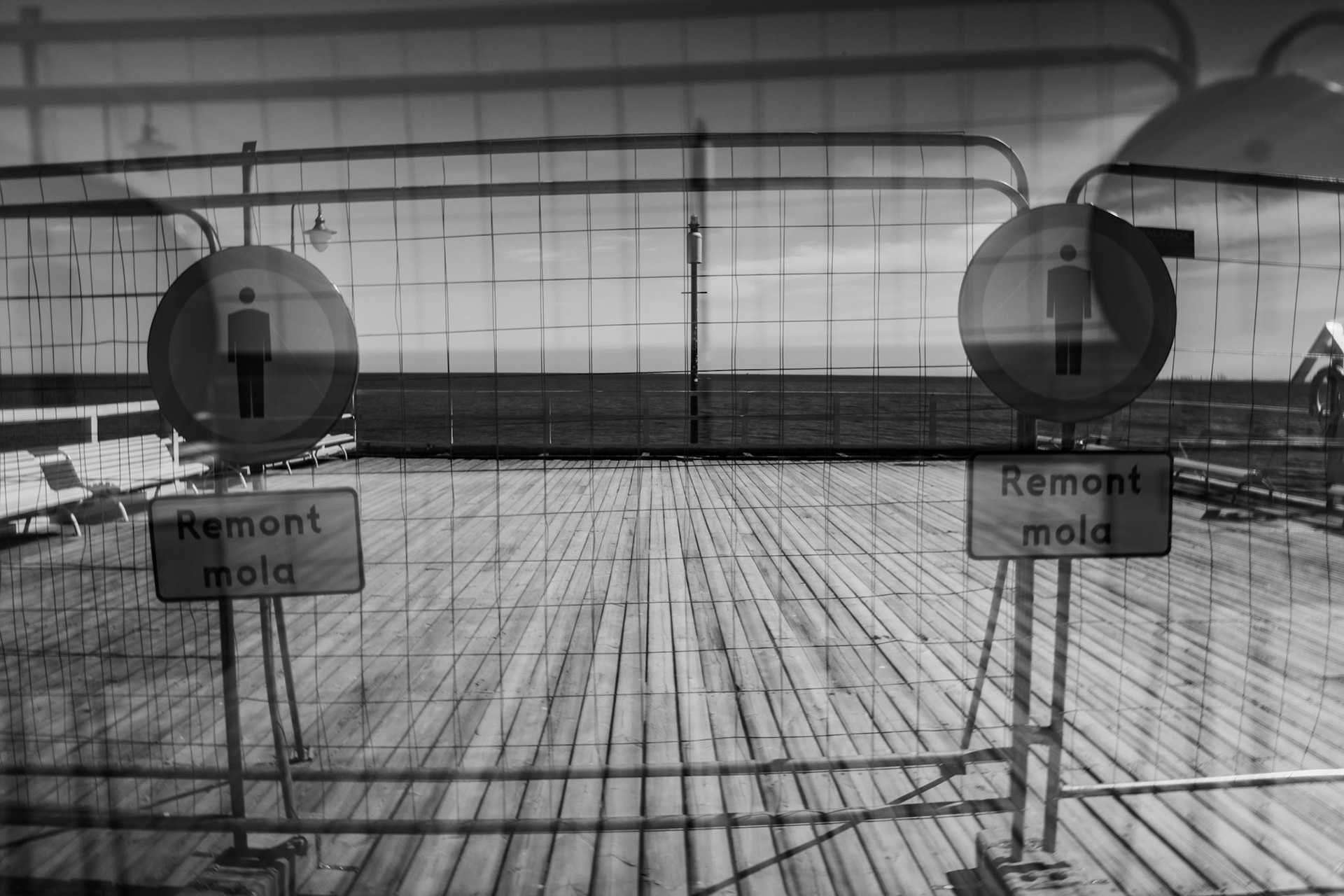 Black-and-white photo of a closed seaside deck with wooden planks, metal fencing, and two signs indicating restricted access, ocean visible in the background.