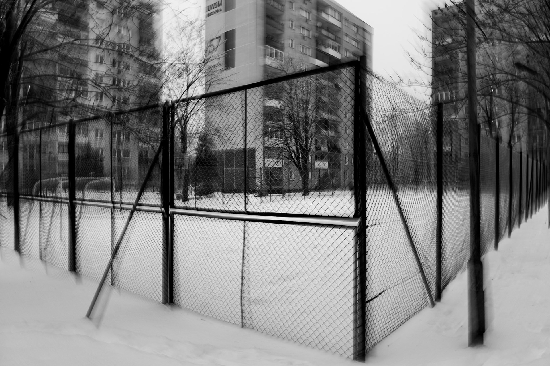 Black and white urban winter scene showing a snow-covered fenced sports court surrounded by tall apartment buildings, bare trees, and motion blur, creating a moody, atmospheric city landscape.