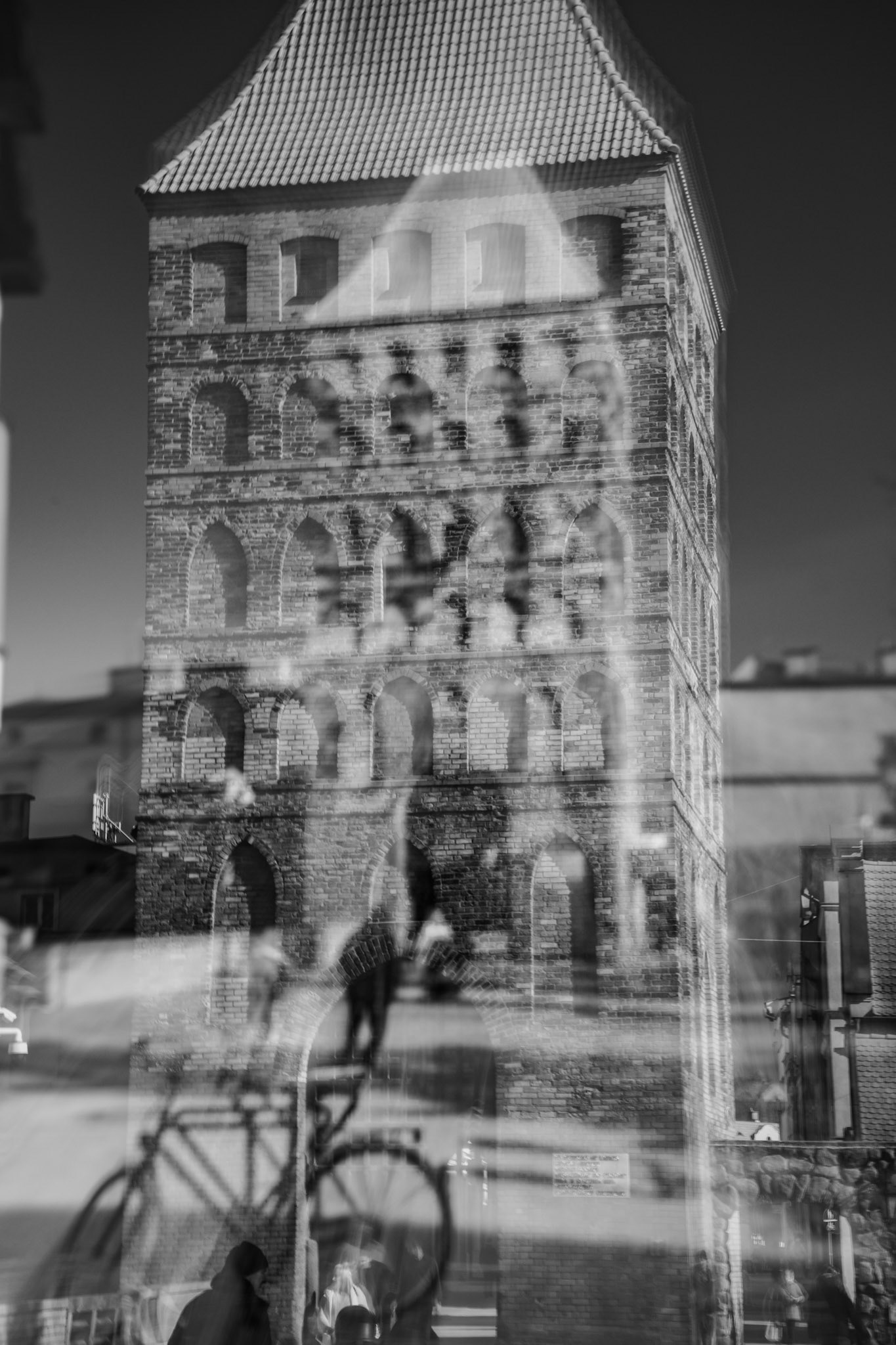A vertical black and white double-exposure of a brick medieval tower. Ghostly silhouettes of a bicycle and pedestrians overlap the ancient architecture.