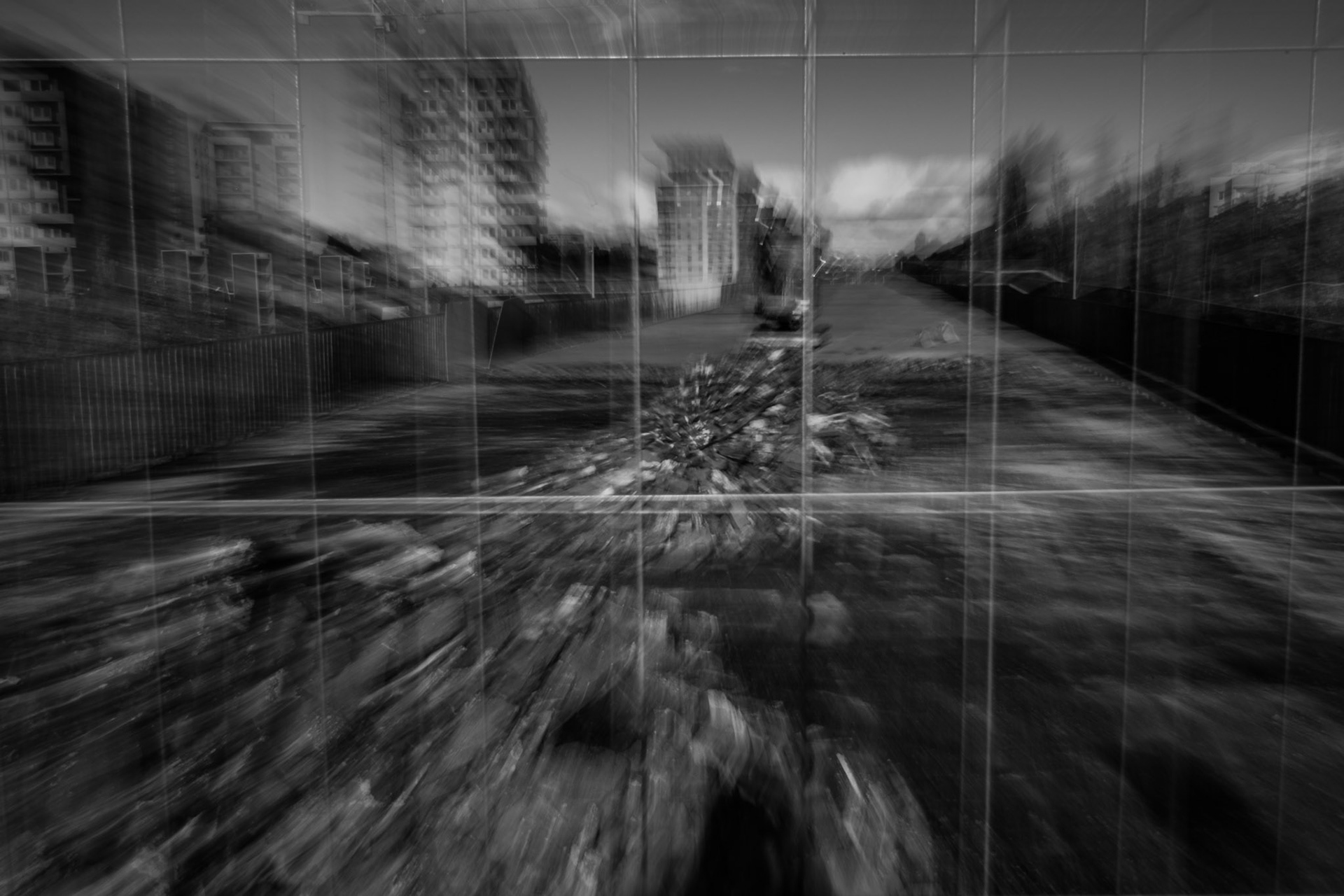 Abstract black and white photograph using the ICM technique, capturing an urban landscape through a fence with motion blur that distorts buildings, rubble, and sky into a dreamlike composition.