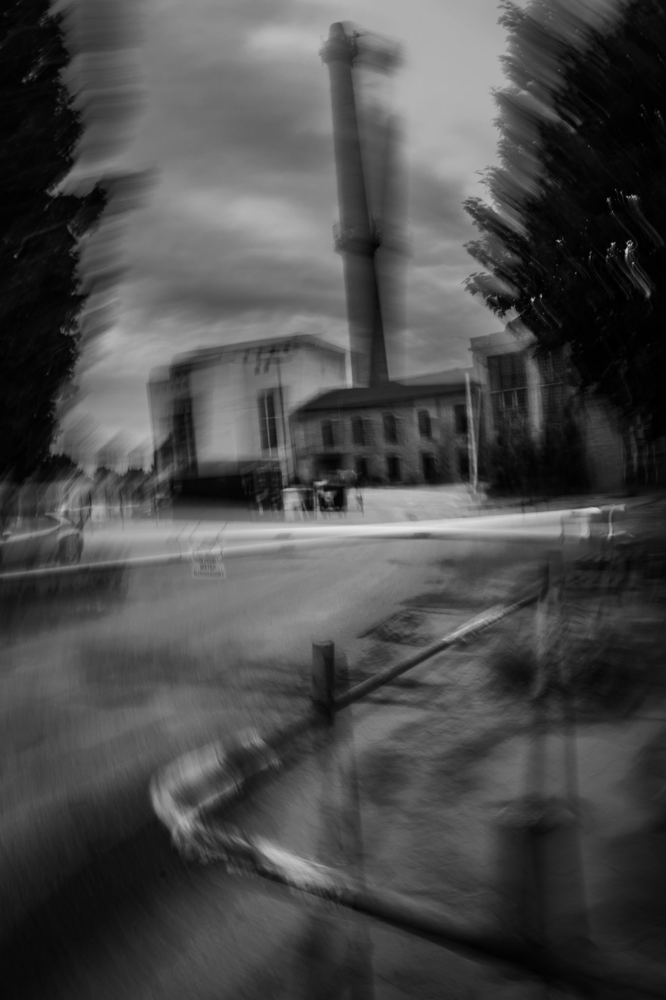 Black-and-white motion-blur photo of an industrial building and tall chimney tower on a rainy, overcast day.