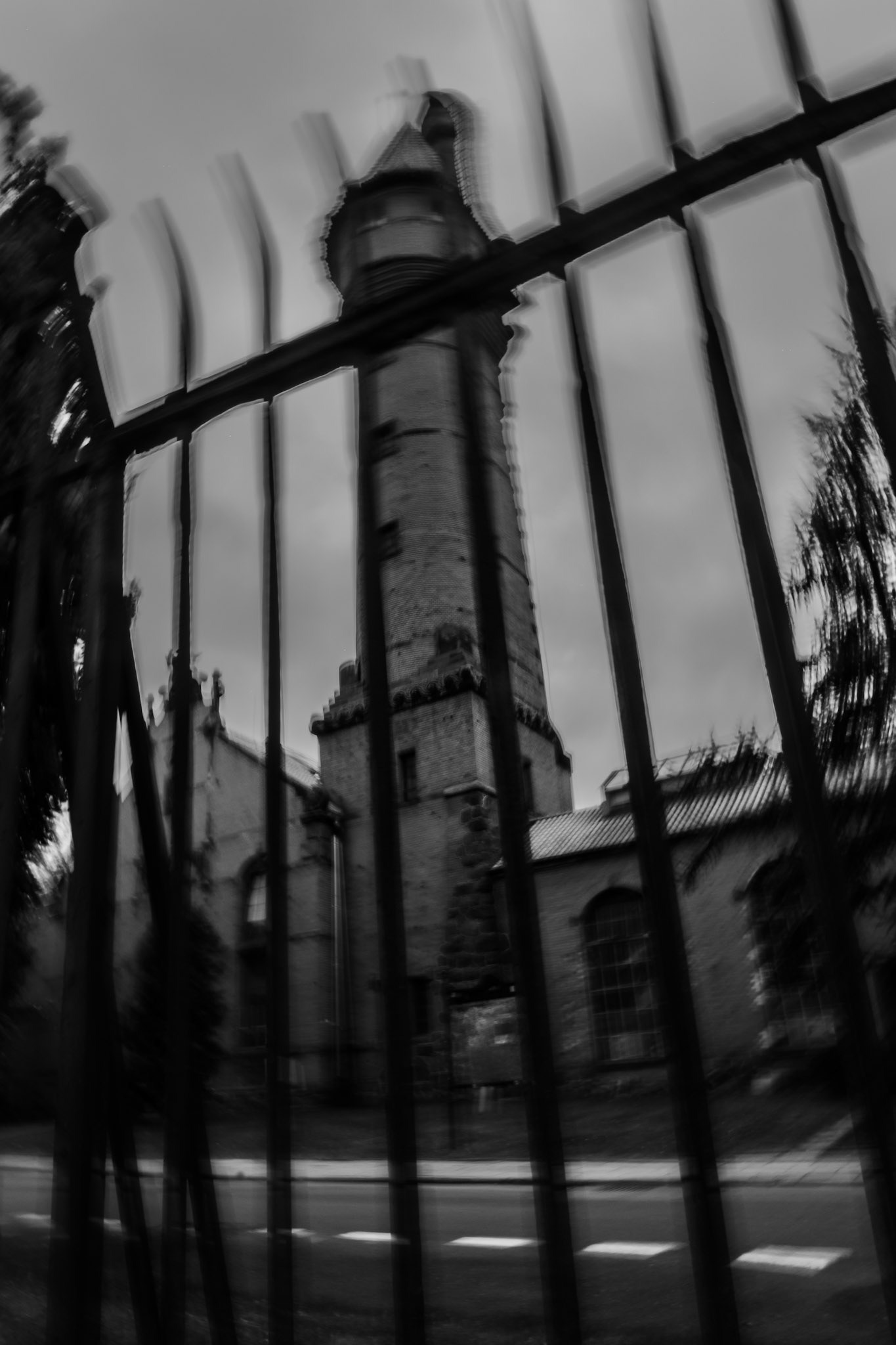 Black and white photo of an old stone tower behind tall iron bars, viewed through a distorted fence, giving a mysterious and eerie atmosphere, made in ICM technique.