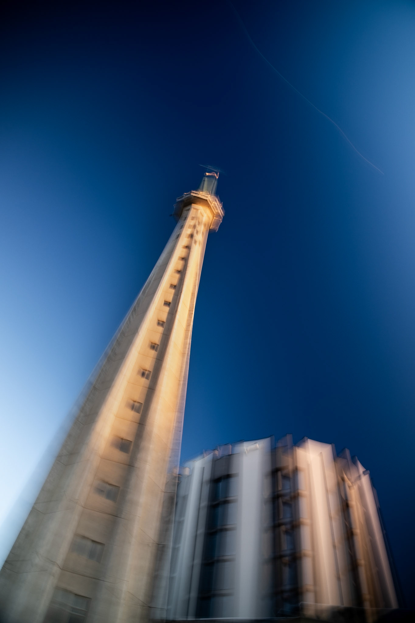 Abstract, motion-blurred photograph of a slender concrete tower rising vertically, captured from a low angle with modern buildings and a dark blue sky.