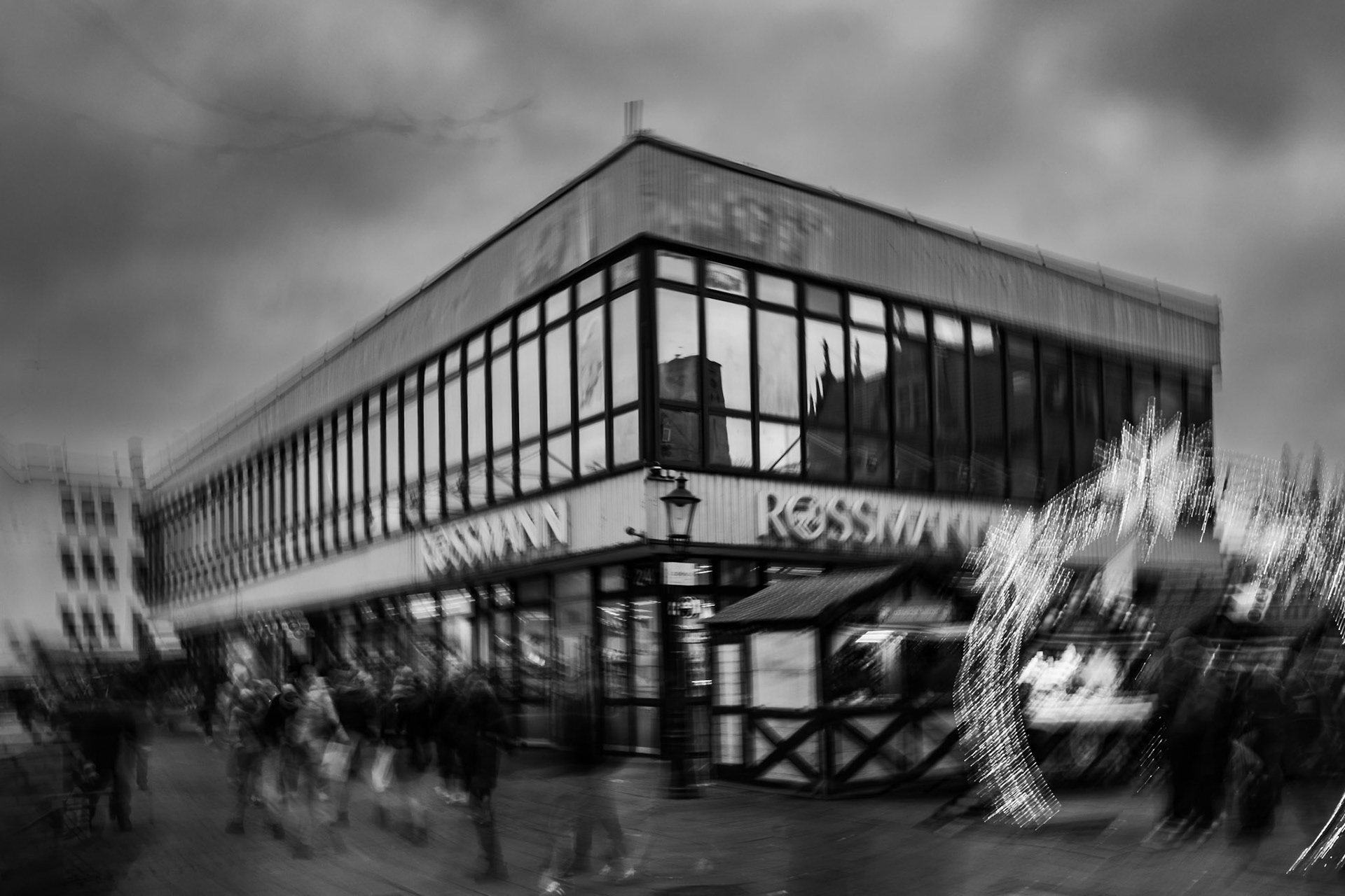Black and white photo of a busy urban street with a modern glass-front building, blurred motion of pedestrians, cloudy sky, and bright festive lights creating a dynamic, atmospheric city scene.