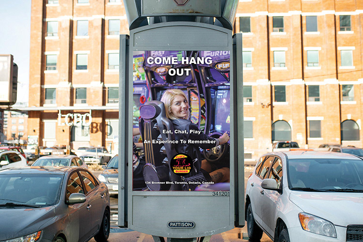In this image is an advertisement that will be placed at the parking lot by the intersection on Front Street West and Spadina Avenue near the Roger Center where it shows a female playing a video game with someone at the restaurant. The words Come Hang Out, Eat, Chat, Play, An Experience To Remember and the location of the restaurant all in a white colour. The Flip'N Your Lid Burger logo is located at the bottom of the advertisement, but above the restaurant's street location in the ad.