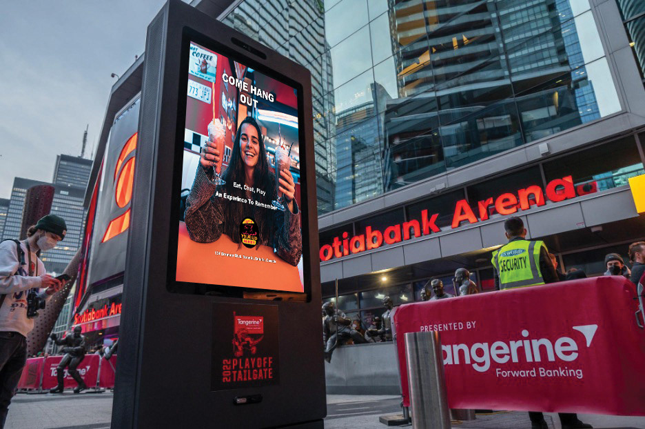 In this image is an advertisement that will be placed near the Soctiabank Arena where in the advertisement is a female that is smiling sitting down at the Flip'N Your Lid Burger retro diner holding two milkshake drinks where one of them is being held on the left hand and the other is being held on the right hand. The words Come Hang Out, Eat, Chat, Play, An Experience To Remember and the location of the restaurant all in a white colour. The Flip'N Your Lid Burger logo is located at the bottom of the advertisement, but above the restaurant's street location in the ad.