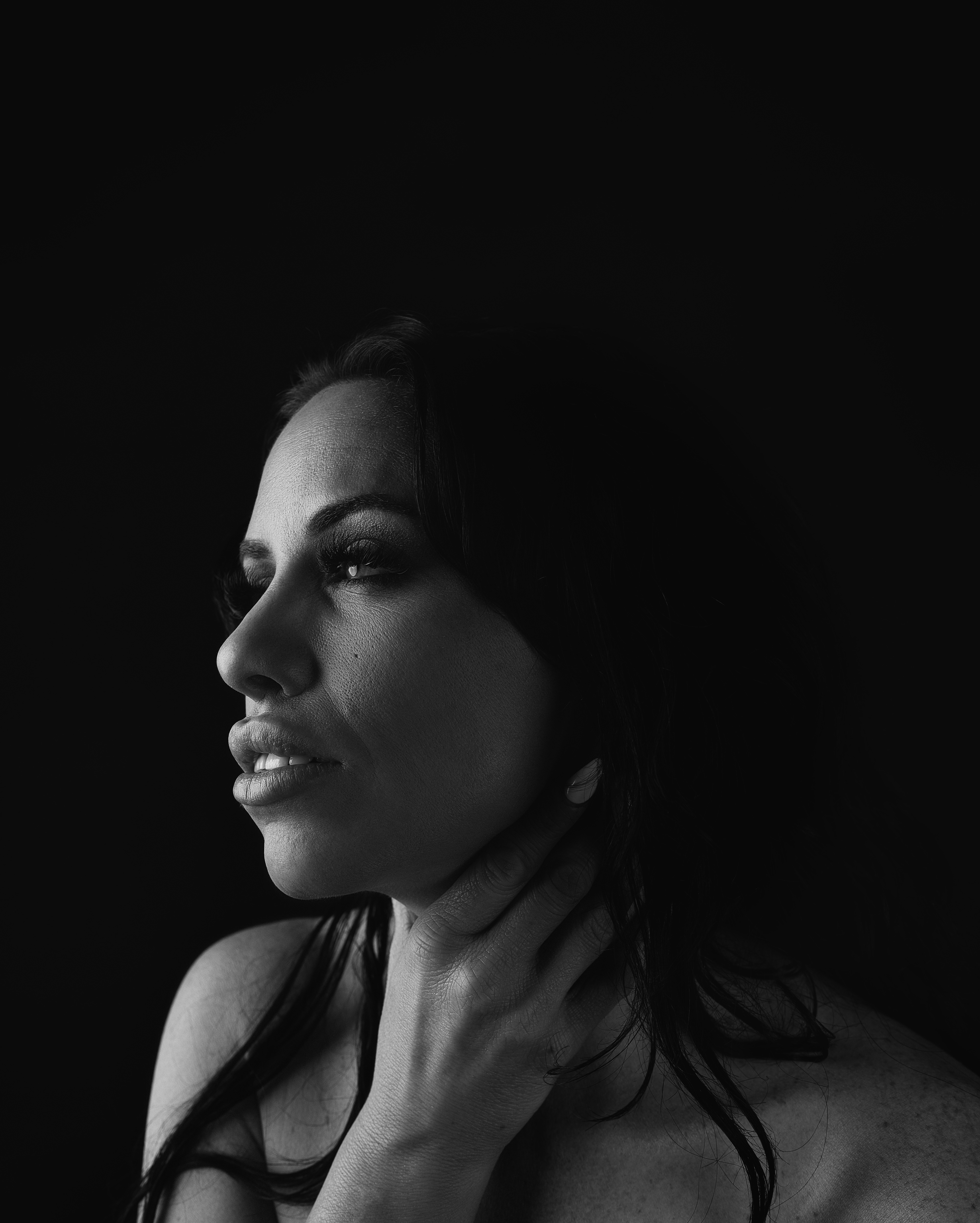 A striking black and white portrait of a woman in Calgary, her gaze intense as she delicately touches her neck. The interplay of light and shadow accentuates her features, evoking a sense of mystery and depth. A captivating artistic expression of emotion and presence