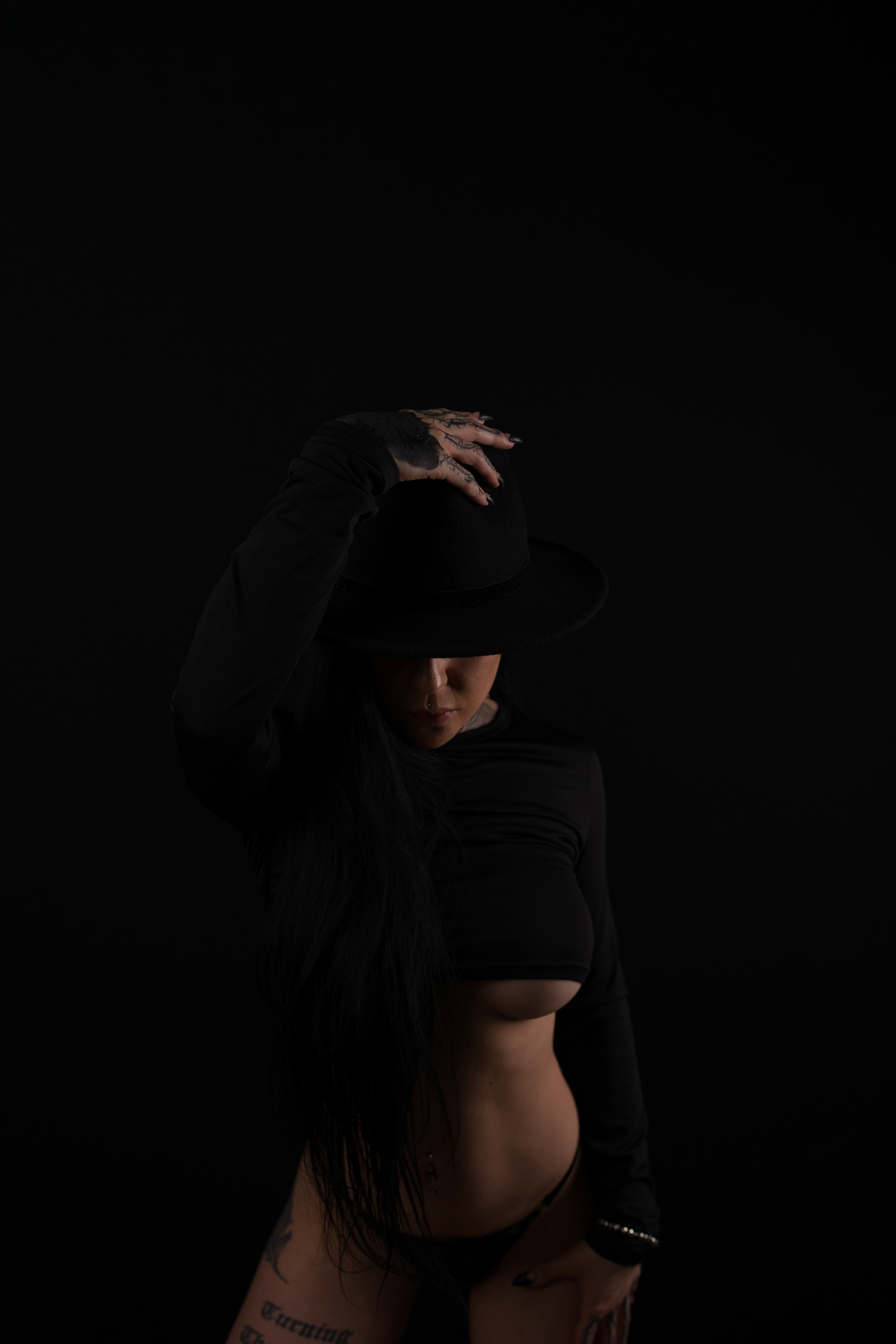 A mysterious and captivating portrait of a woman in Calgary, draped in darkness with only the subtle glow of light revealing her presence. Dressed in all black, she tips her wide-brimmed hat, concealing part of her face, while long, flowing hair cascades over her shoulder. The interplay of shadows and form creates a bold yet intimate moment, embodying strength, allure, and artistic elegance