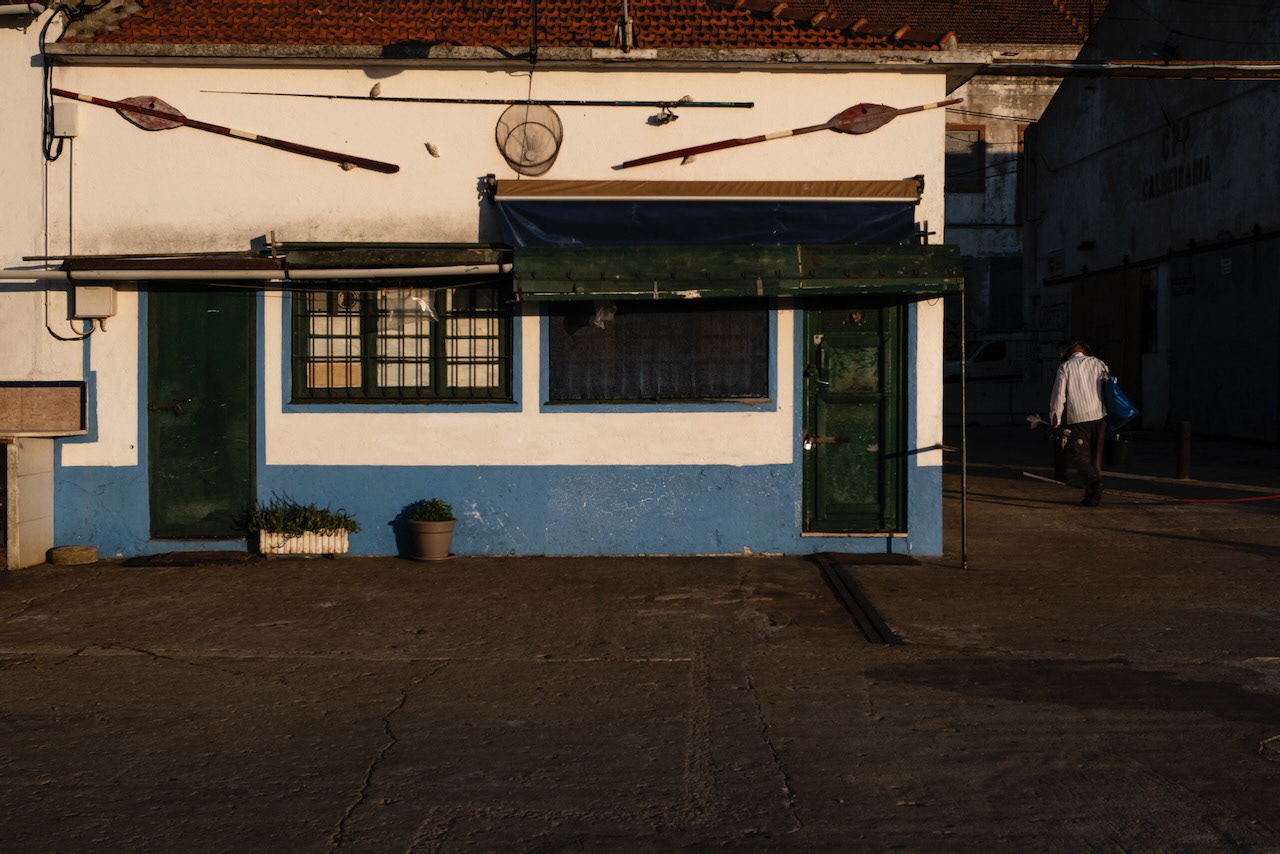 Under the light of the setting sun, the wall of a fishermen's warehouse painted in white and blue and a fisherman in a white shirt walking away.
