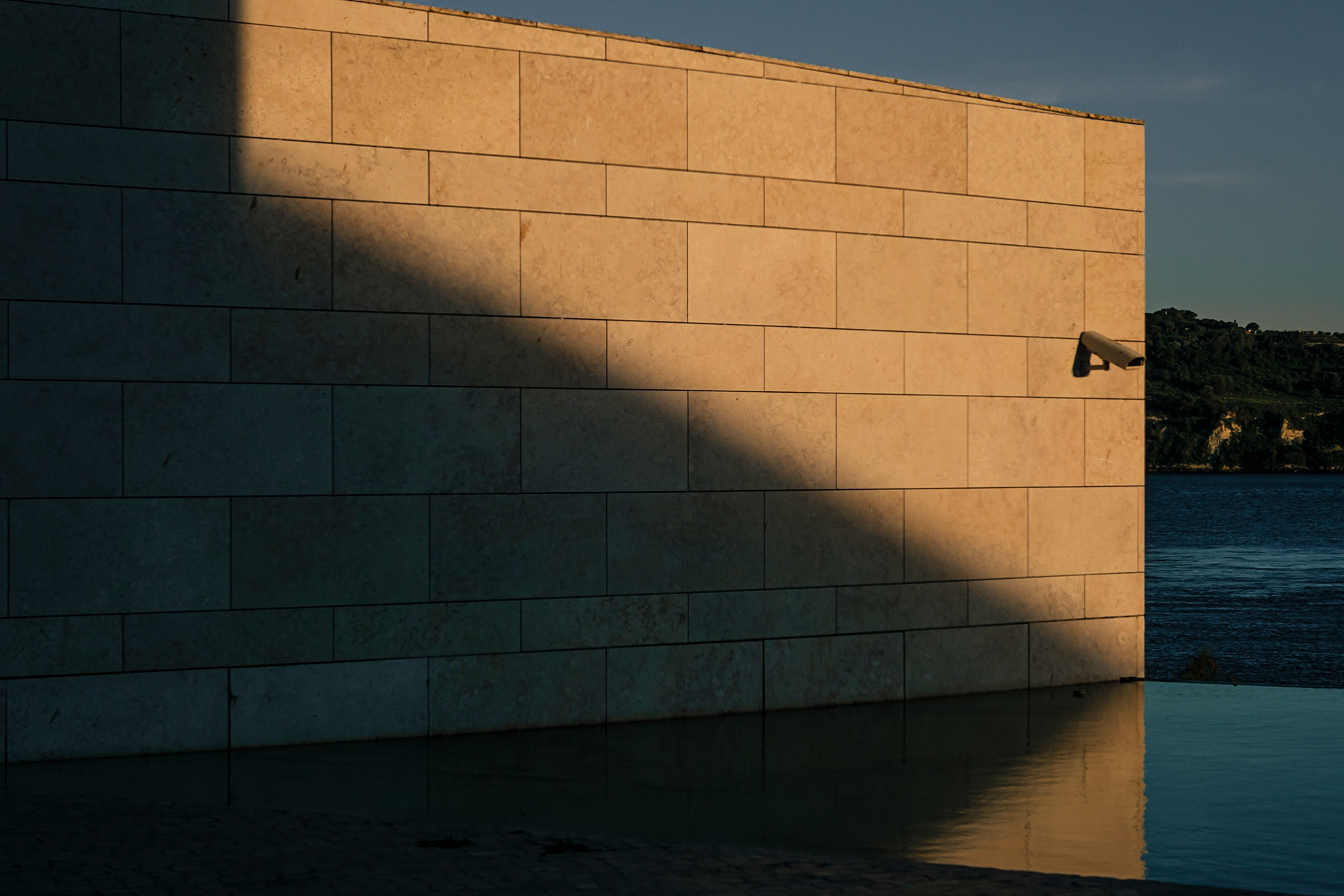 The light of the setting sun falls down on a stonewall that is reflected on a pool. A surveillance camera is in the light. An oblique shadow darkens part of the wall.