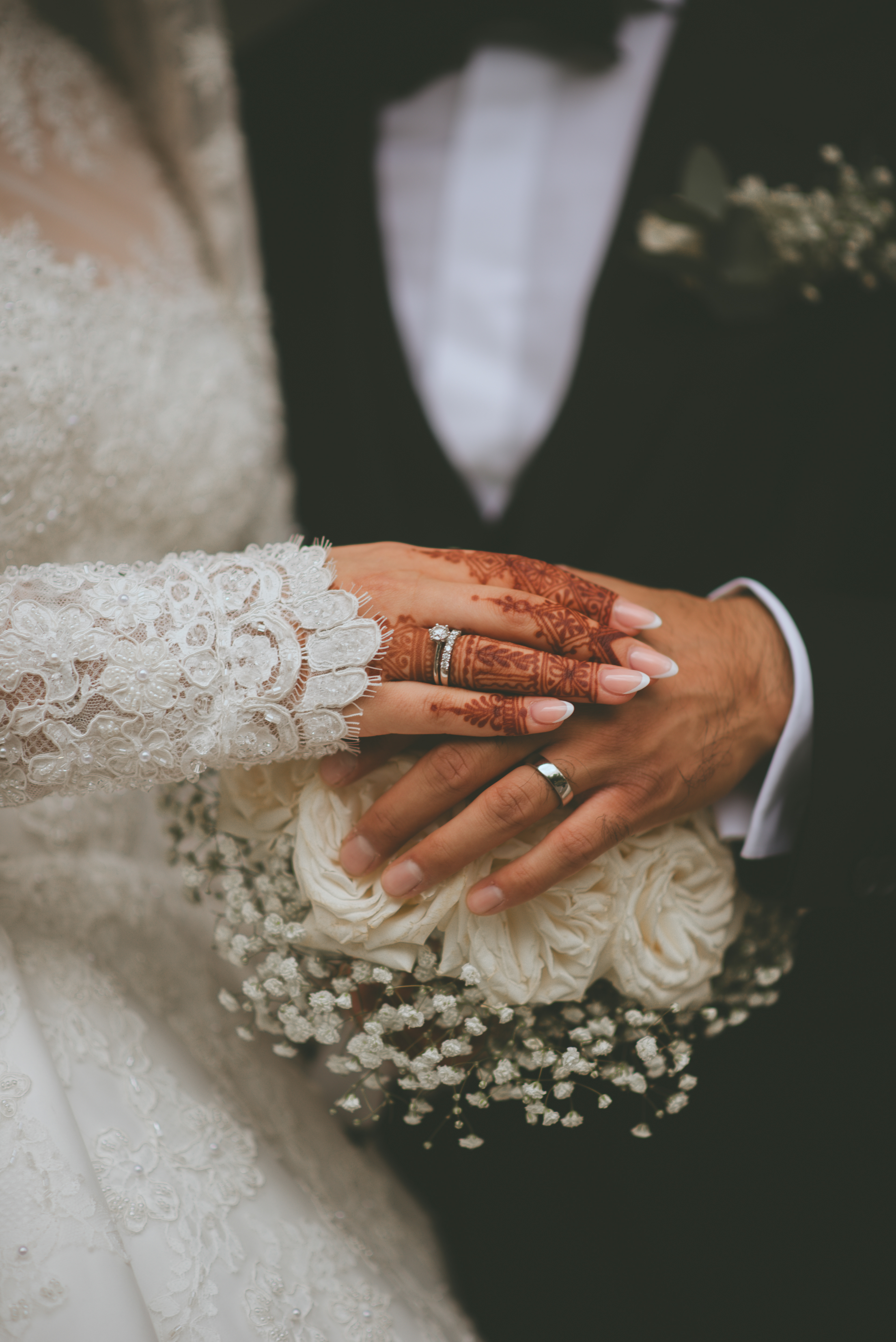 Das Bild zeigt eine Nahaufnahme der Hände eines Brautpaares, die auf einem weißen Rosenstrauß liegen. Die Hände der Braut sind mit aufwendigen Henna-Mustern und spitzenverzierten Ärmeln geschmückt. An beiden Ringfingern sind Eheringe zu sehen.