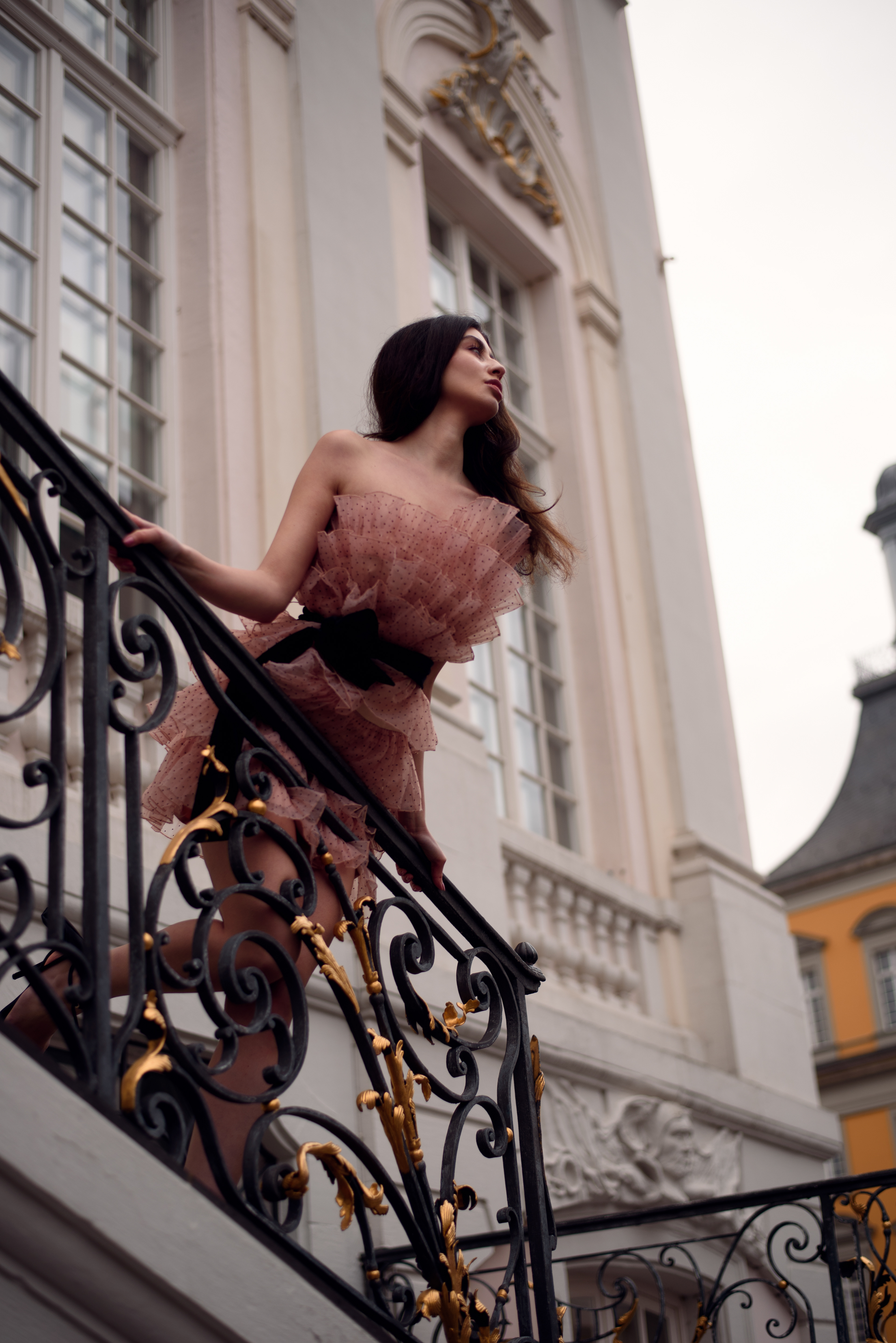Ein Porträtfoto von einer Frau, die auf einer kunstvollen Treppe steht. Sie trägt ein kurzes, mehrlagiges Kleid in einem rosafarbenen Ton und einen schwarzen Gürtel. Im Hintergrund ist ein helles, klassisches Gebäude mit großen Fenstern zu sehen.