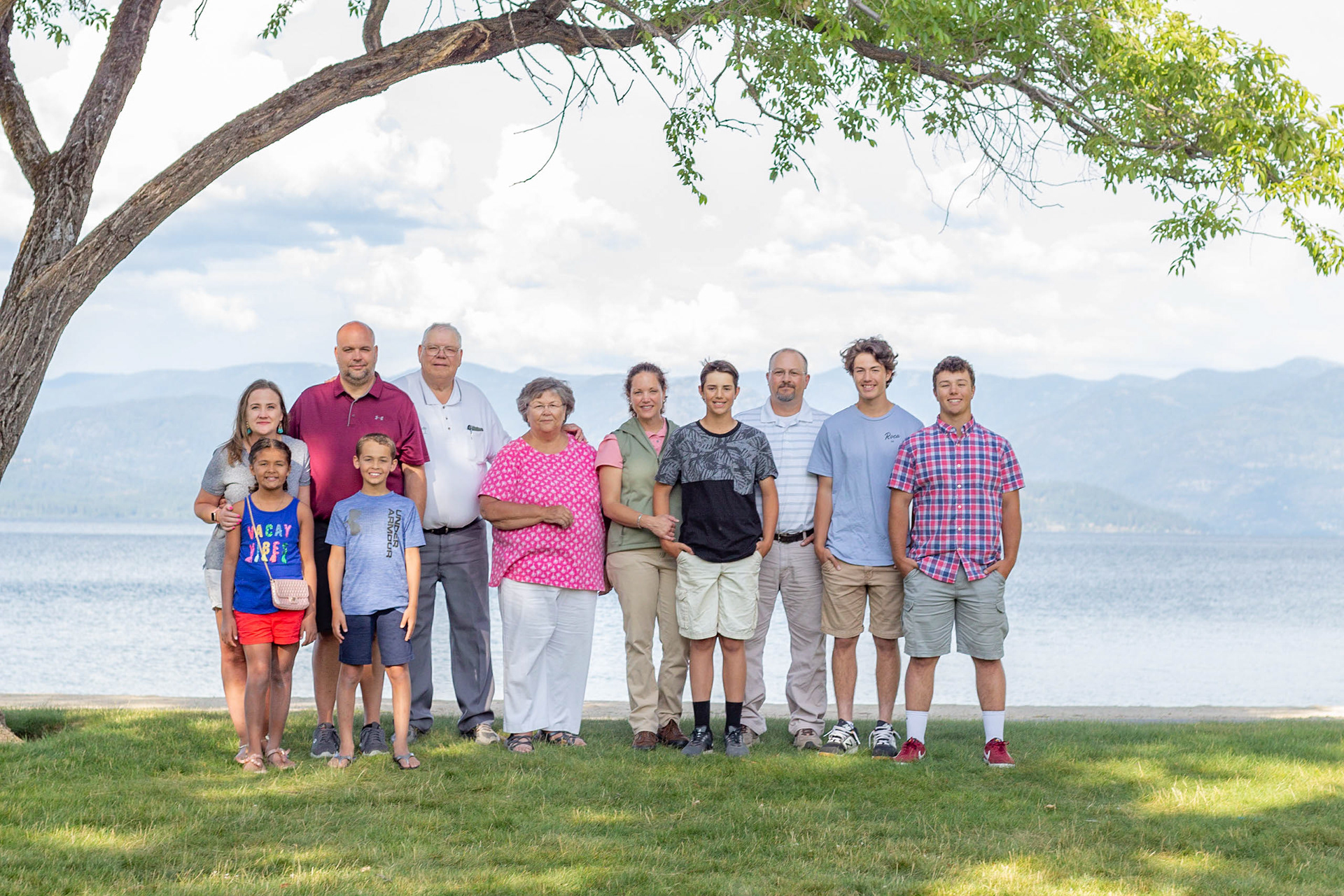 Multi-generational family portrait outdoors under a tree by Lake Pend Oreille in Sandpoint, North Idaho, with mountains and water in the background during a summer family photography session.