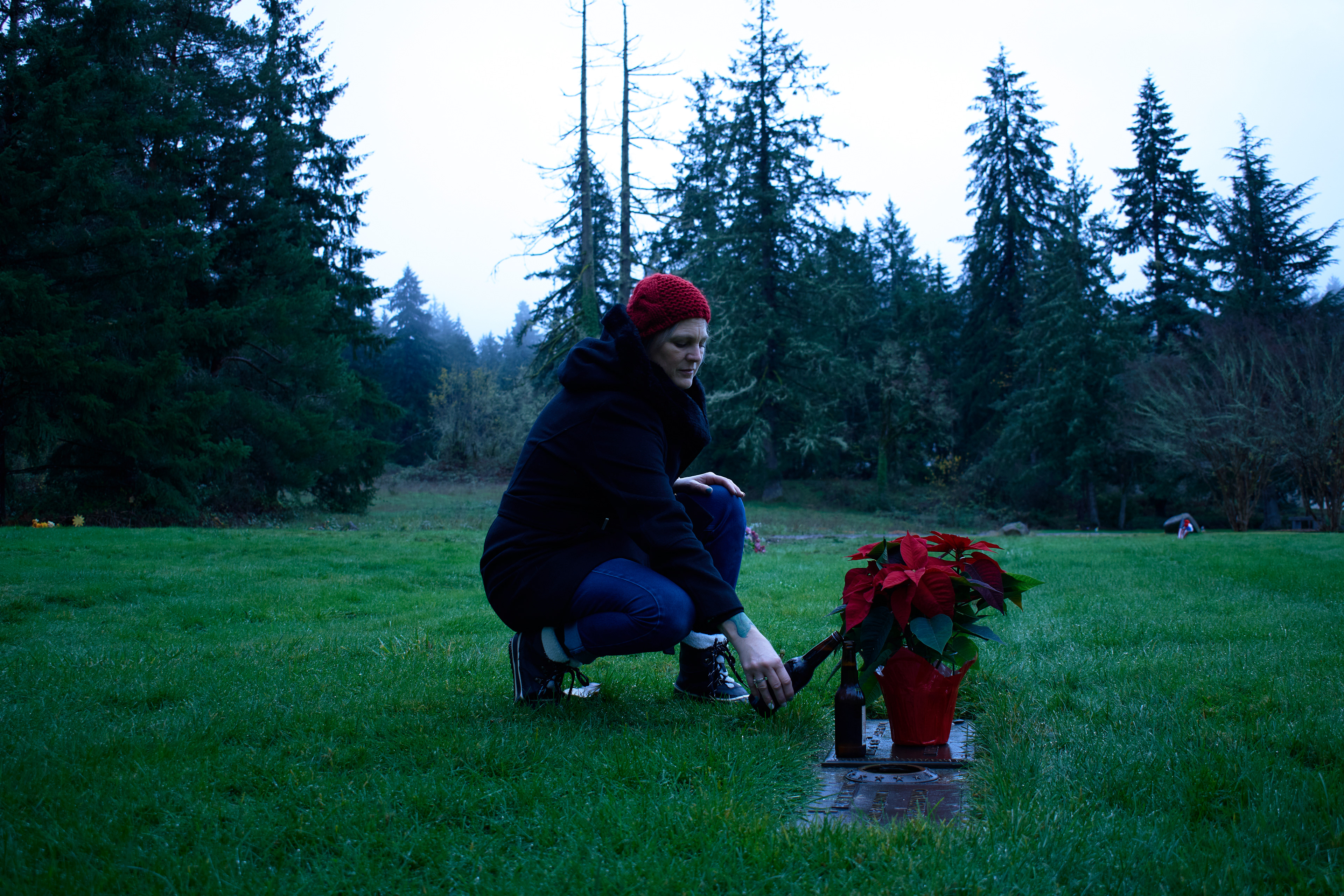 Woman in a red beanie kneeling by a grave with a beer in her hand, clinking it against an open bottle on top of the grave. 
