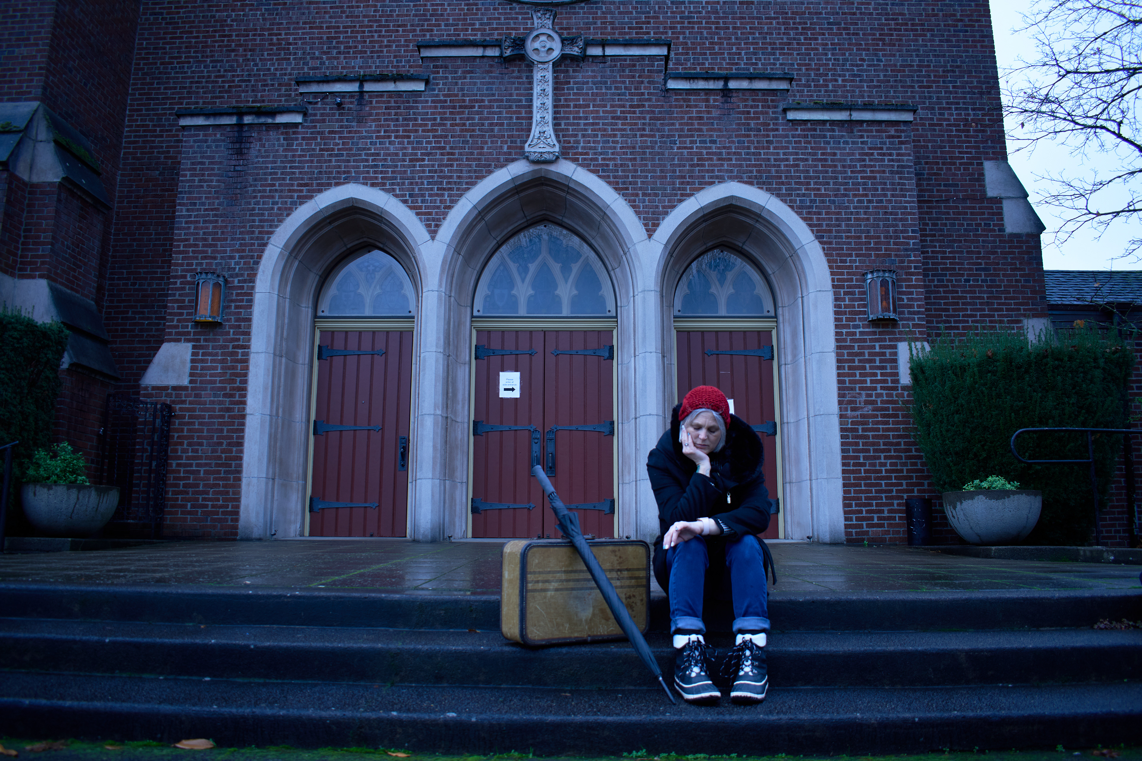 Woman in a red beanie sitting outside of a Catholic Church looking at her watch.