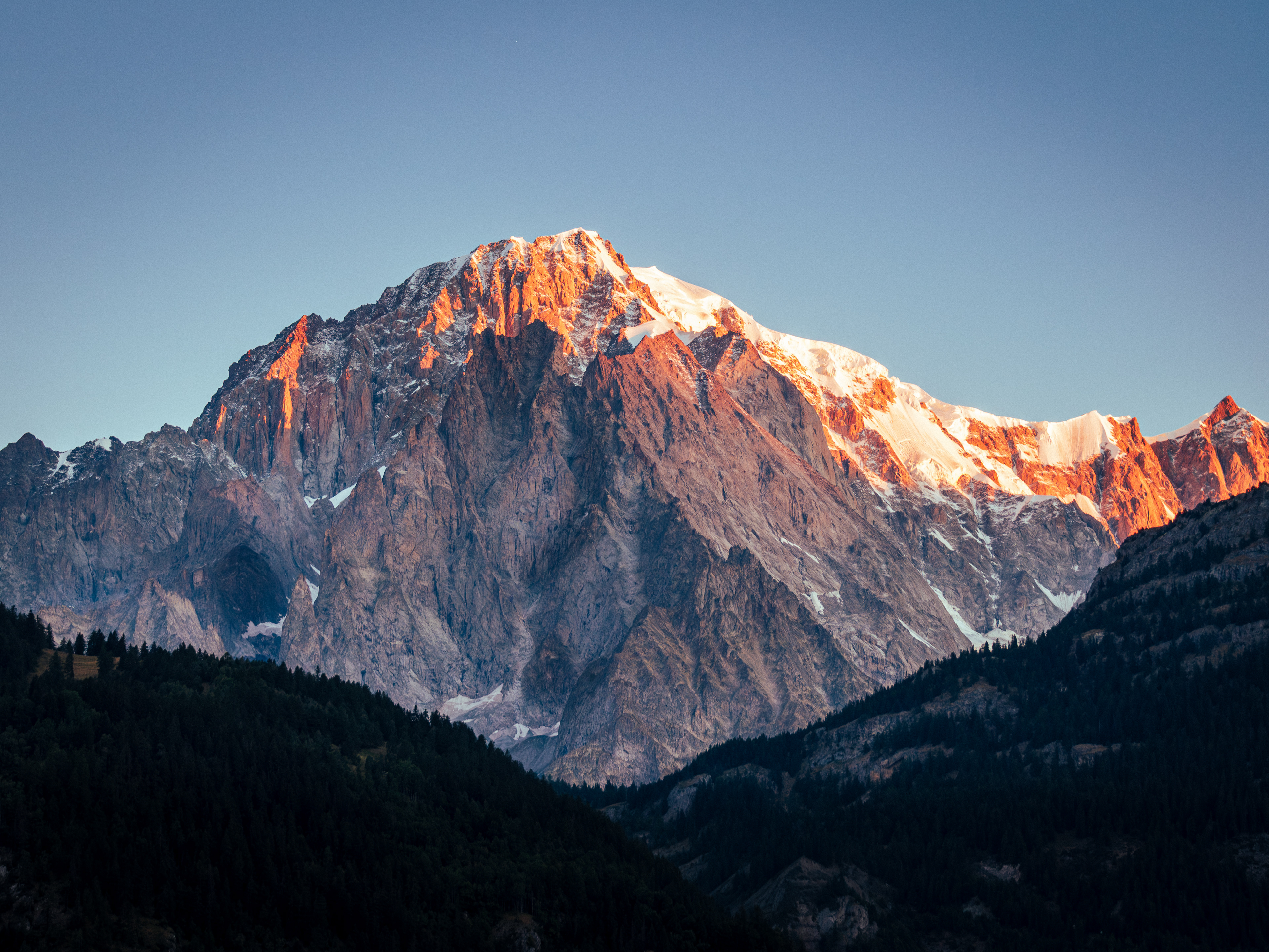 ‘The Vast, Quiet Certainty’: From the Transition Journeys series, a massive, rugged mountain face captured in the warm glow of alpenglow, standing in sharp contrast against a dark, forested foreground and a clear twilight sky.