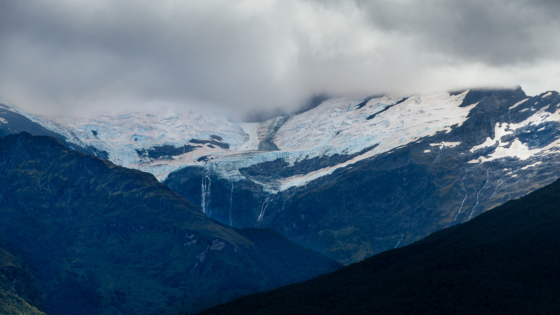 Glacier under Clouds’: Part of the New Zealand series, showing a massive, fractured blue glacier nestled between dark mountain ridges, with several waterfalls cascading down the rock face into a hidden valley.
