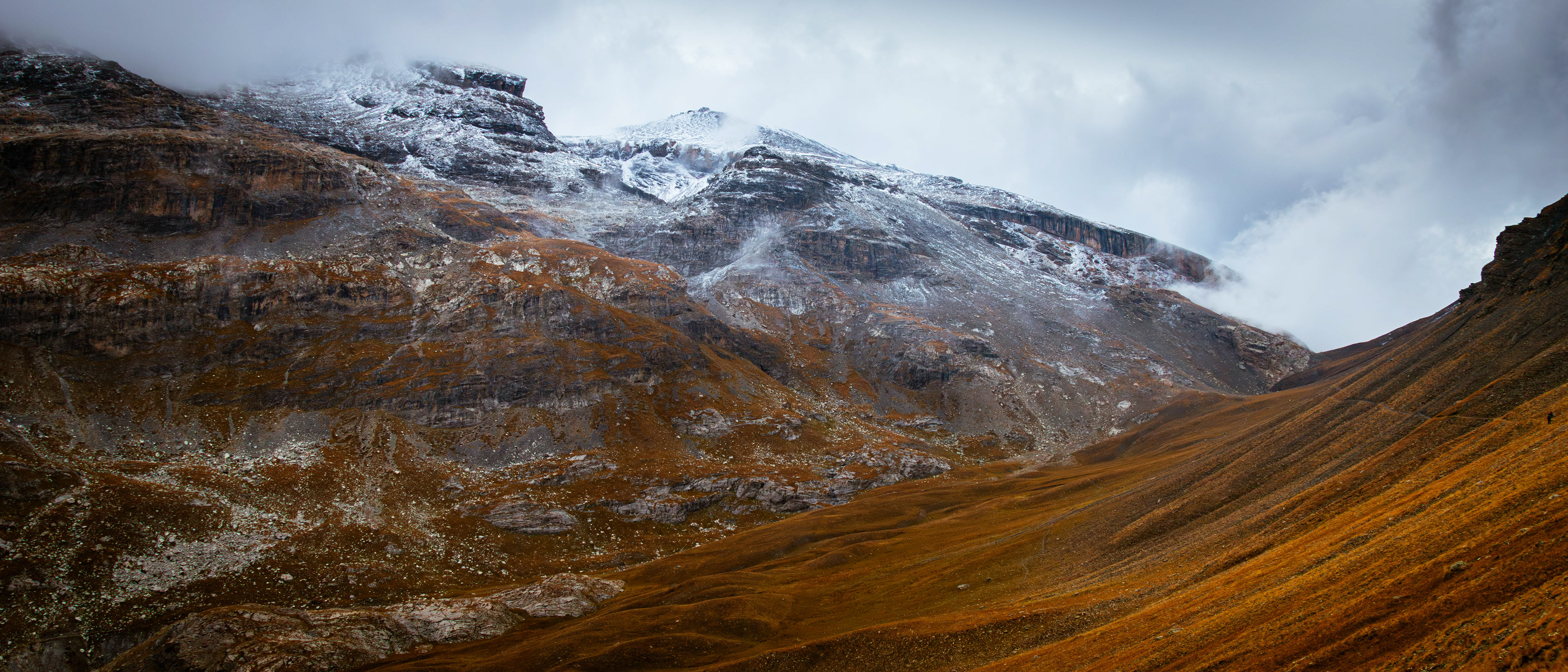 ‘Autumn Snow Clouds’: A wide-angle landscape shot from the Alpine Moods series, showing a vast, golden-brown alpine valley beneath heavy, leaden clouds and distant snow-covered ridges.
