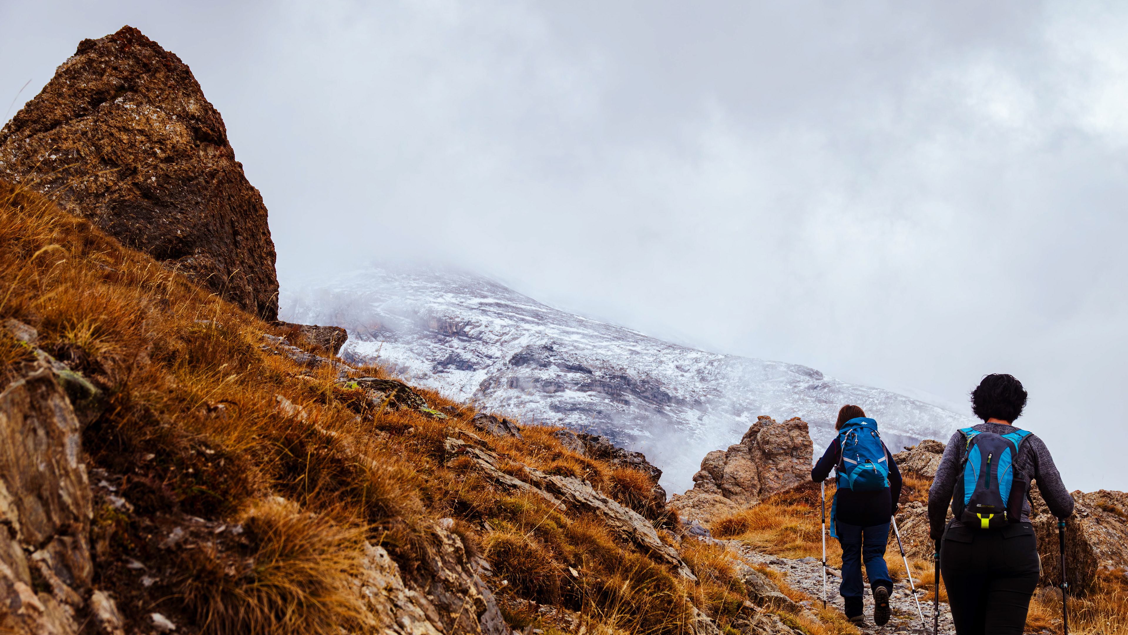 ‘Mountains Revealed’: From the Alpine Moods series, two hikers ascend a steep, rocky trail toward a formidable, snow-streaked peak that is partially veiled by thick, white mountain mist.