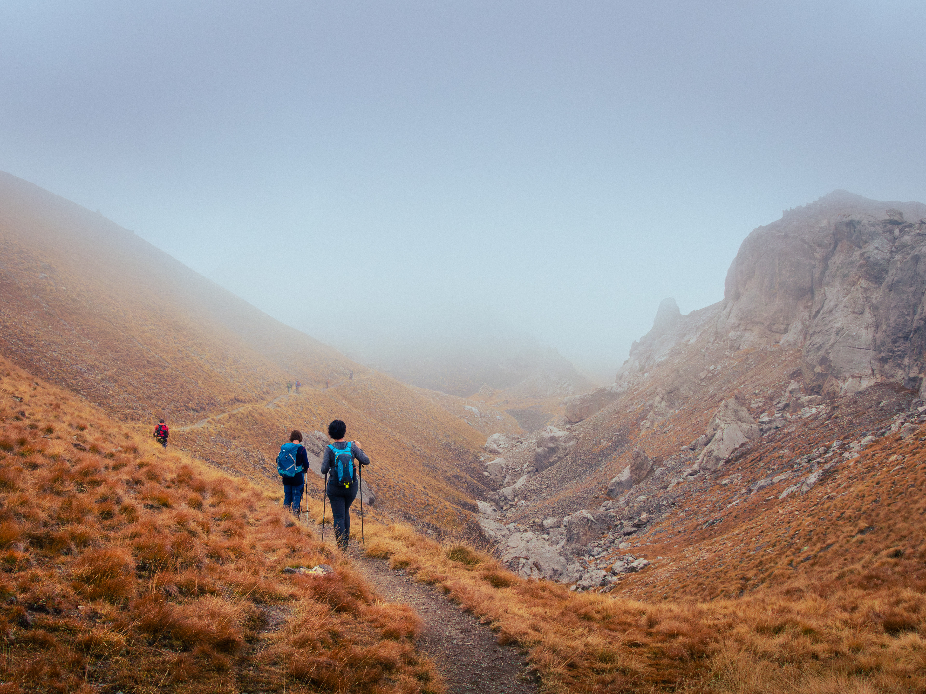‘Quest into the Unknown’: From the Transition Journeys series, two hikers follow a dirt trail into a mountain valley obscured by dense white fog, surrounded by slopes of golden-brown autumn grass.