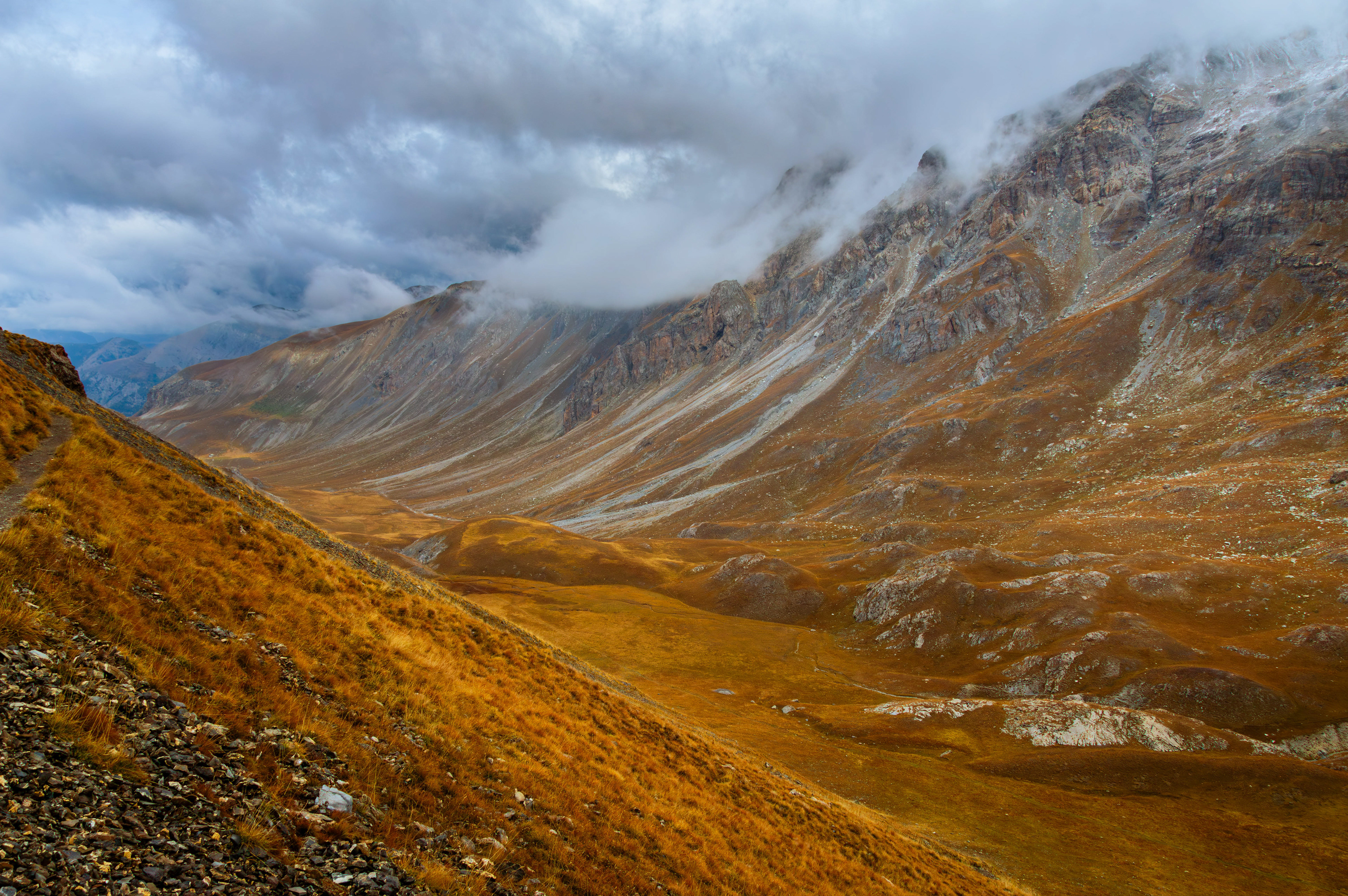 ‘Down the Valley’: Part of the Alpine Moods series, featuring a dramatic view down a sweeping, golden-hued valley in the French Alps, with swirling clouds clinging to the steep, rocky slopes.