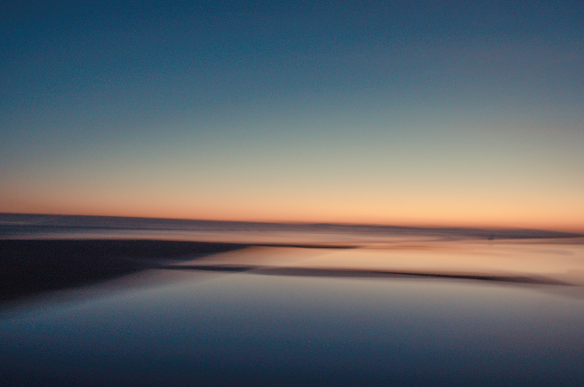 West Kirby marine lake in the evening, the sky is darkening to deep blue and there is a blush of orange and gold along the horizon. The tide is out and the sunset is reflected in the wet sand. The marine lake is quiet, the water is still, the water reflects the deep blue of the sky and the golden shades of the sunset. The image is taken using intentional camera movement  to an over all soft effect.