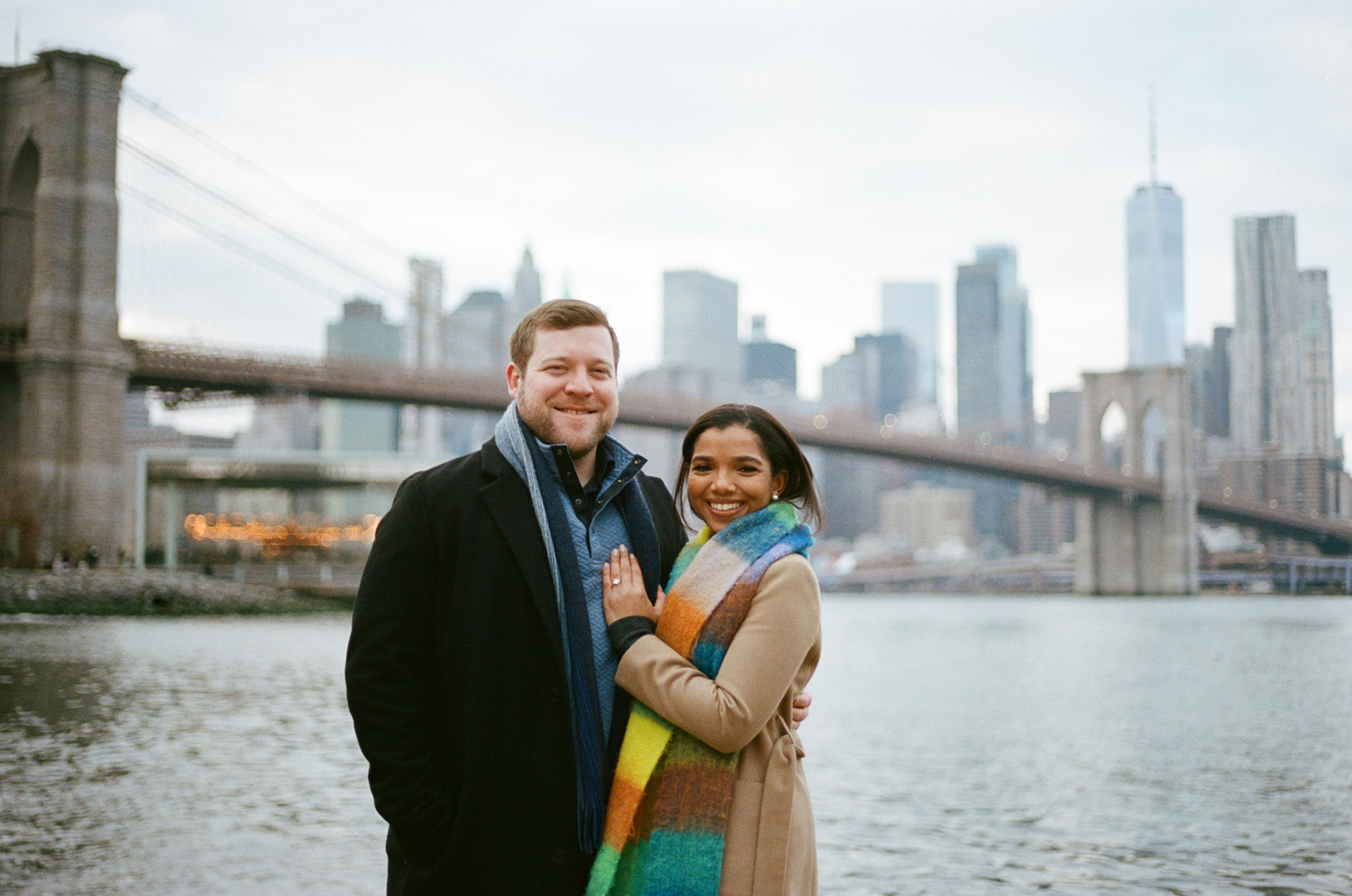 35mm film portrait of a couple with the NYC skyline and Brooklyn Bridge in the background, with a soft vintage aesthetic.