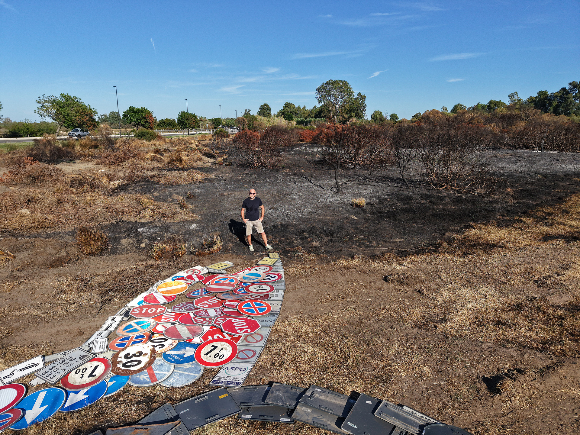 Artist Scoobafish standing beside the road-sign flame of IT TAKES SO LITTLE., with charred ground and burnt shrubs behind him in San Teodoro, Sardinia.