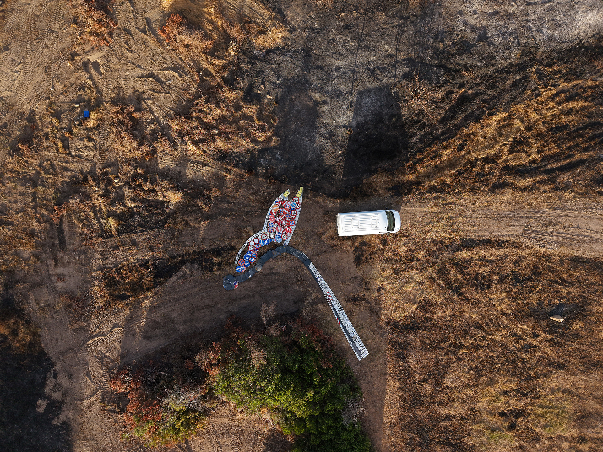 Aerial view of IT TAKES SO LITTLE. by Scoobafish, a half-burnt match made from reclaimed road signs on fire-scarred ground, with a white van beside it providing a sense of scale.