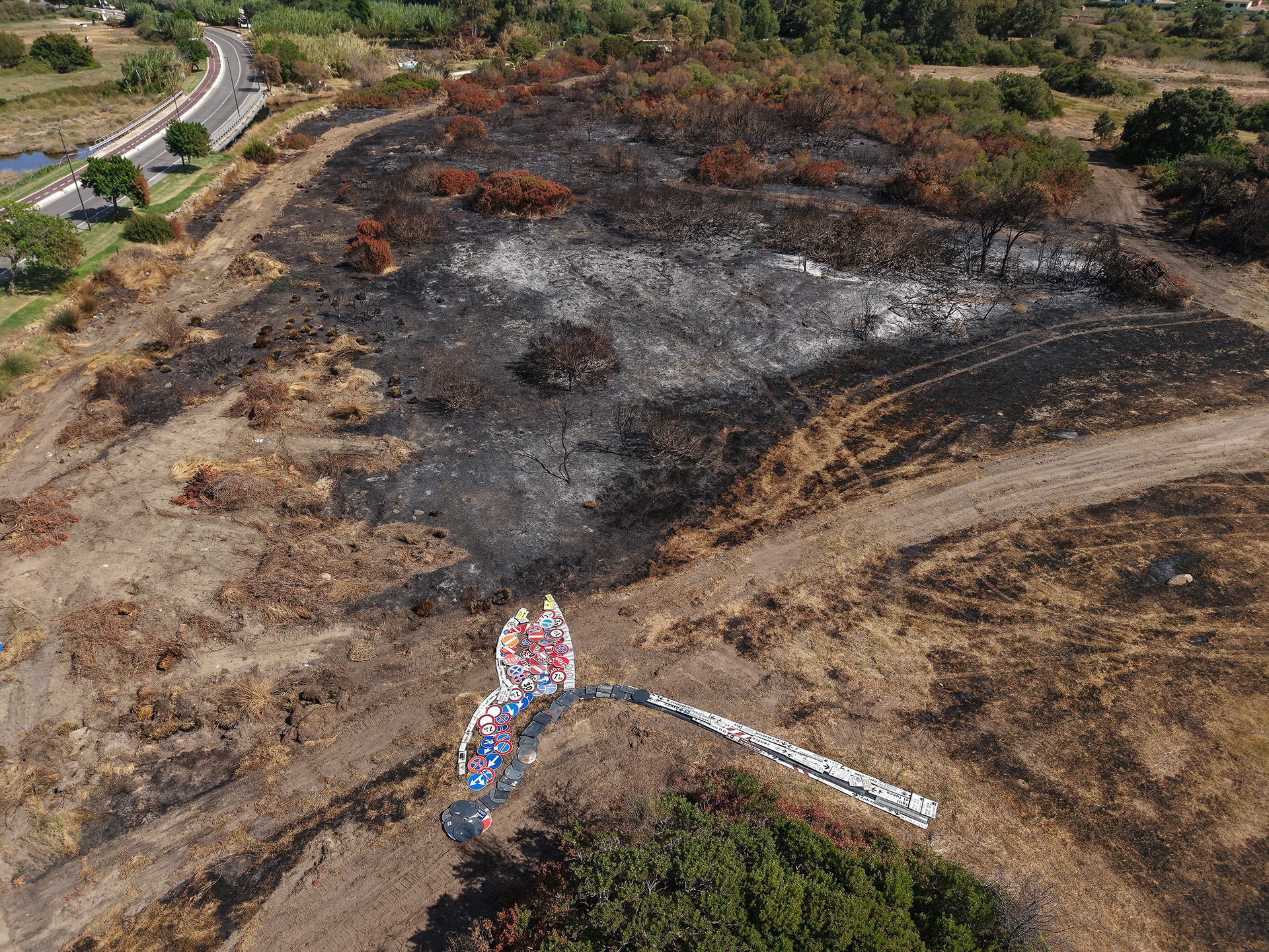Oblique aerial view of IT TAKES SO LITTLE. by Scoobafish: a burning match assembled from reclaimed road signs below an expanse of fire-scarred ground in San Teodoro, Sardinia.