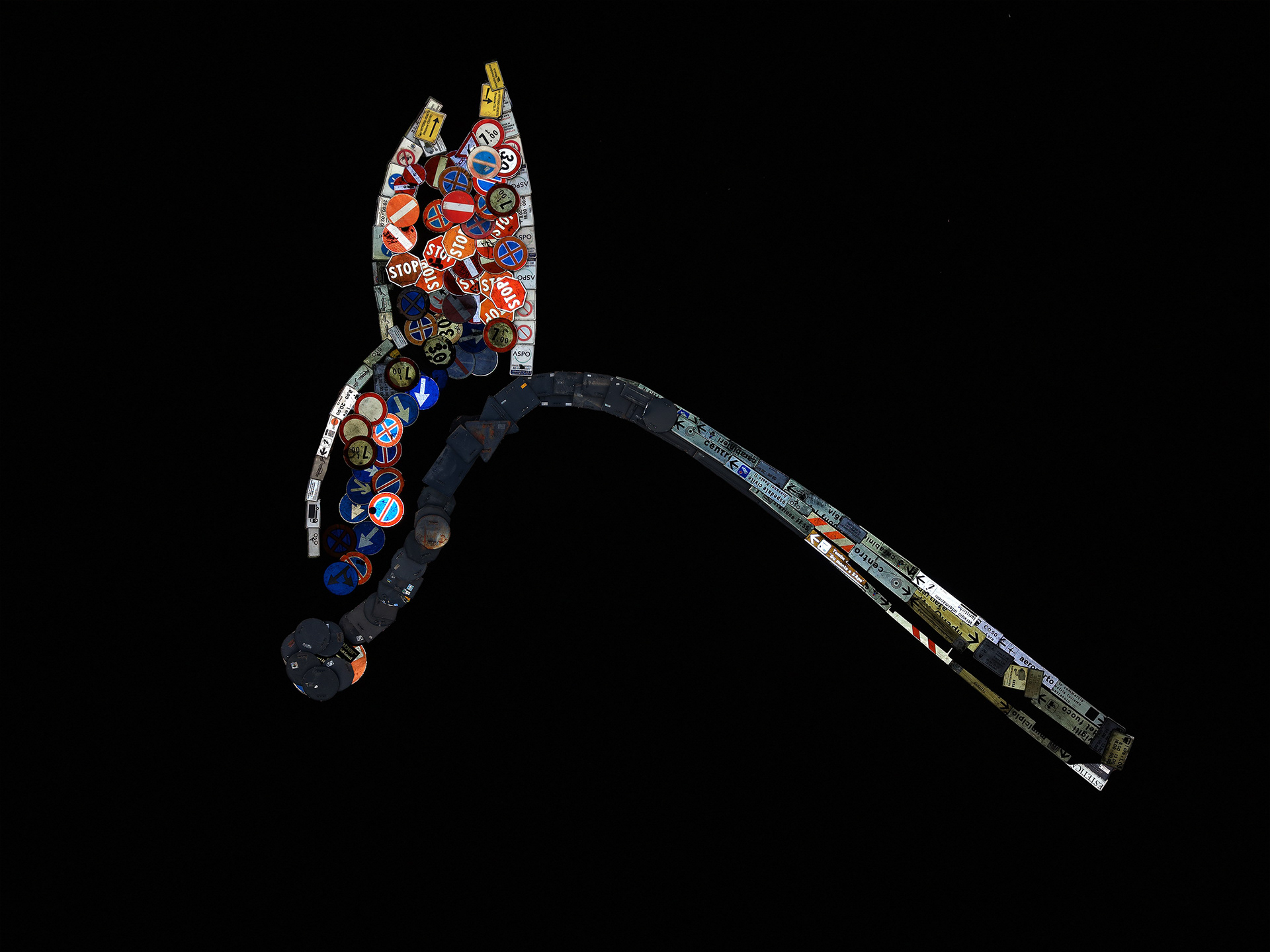 Night view of IT TAKES SO LITTLE., with reflective road-sign faces defining the flame and slender stem, while grey metal backs trace the curved, charred section against the dark ground.