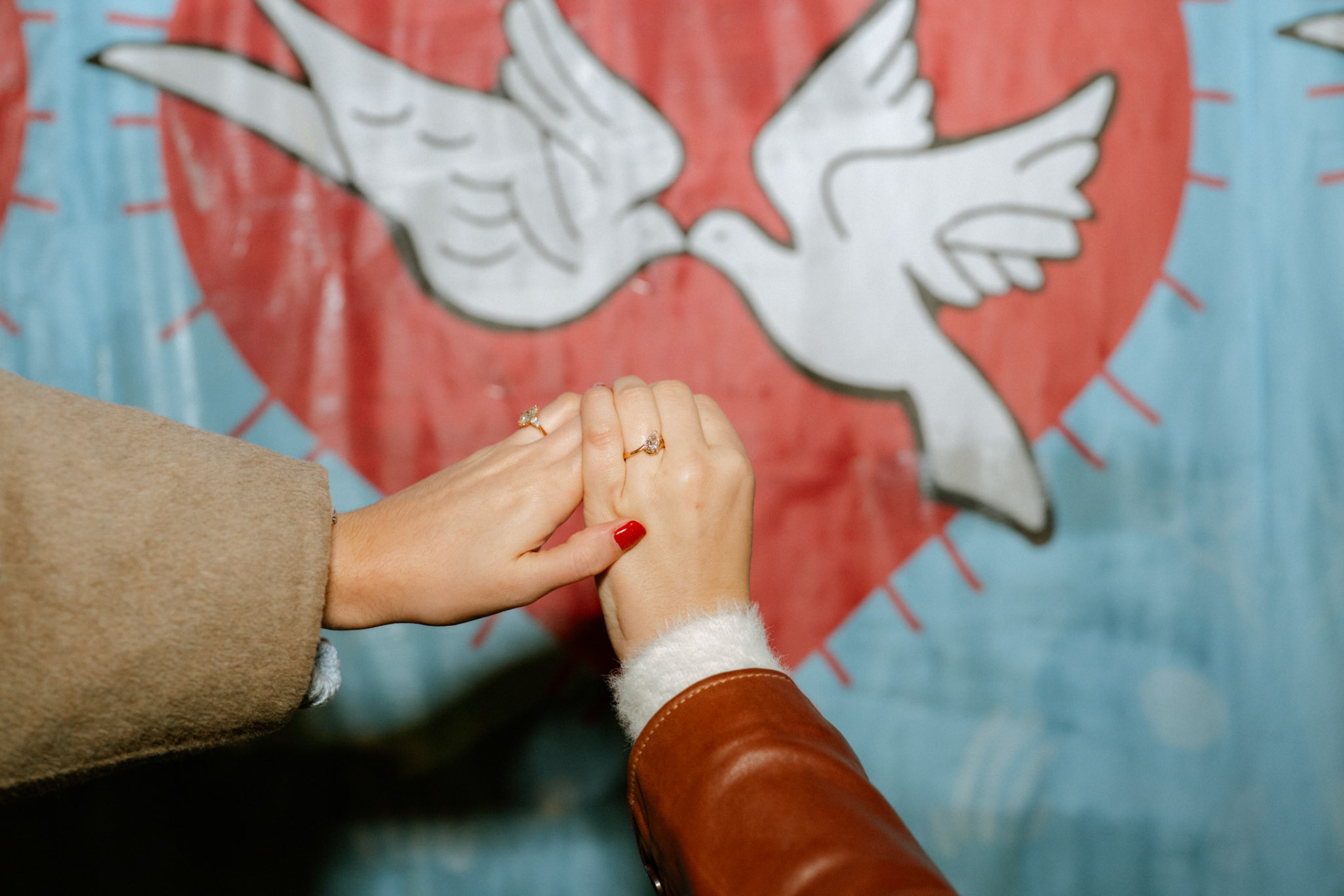 Newly engaged couple holds hands in front of a poster with a heart and doves