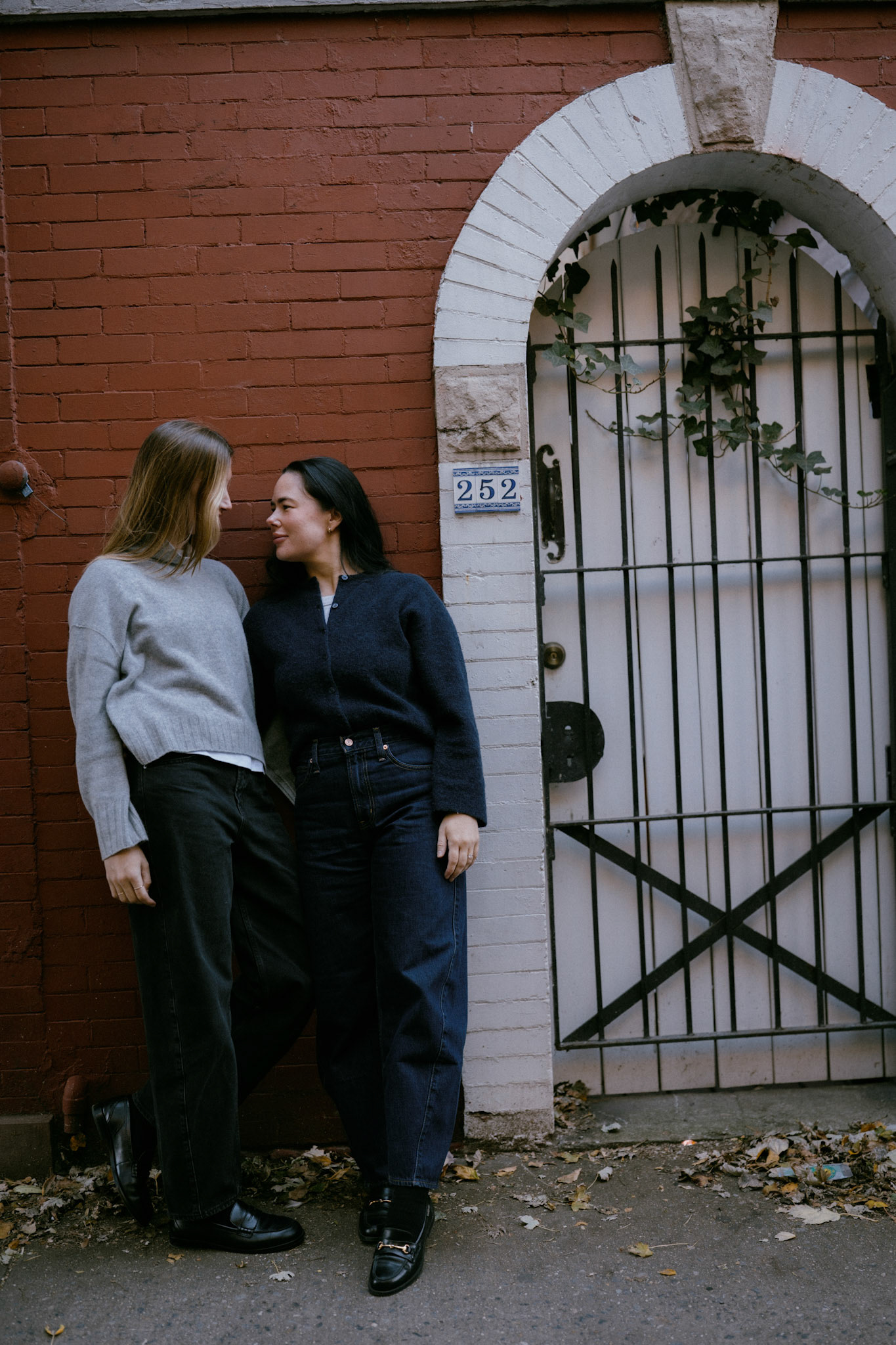 Two women lean against a brick wall.
