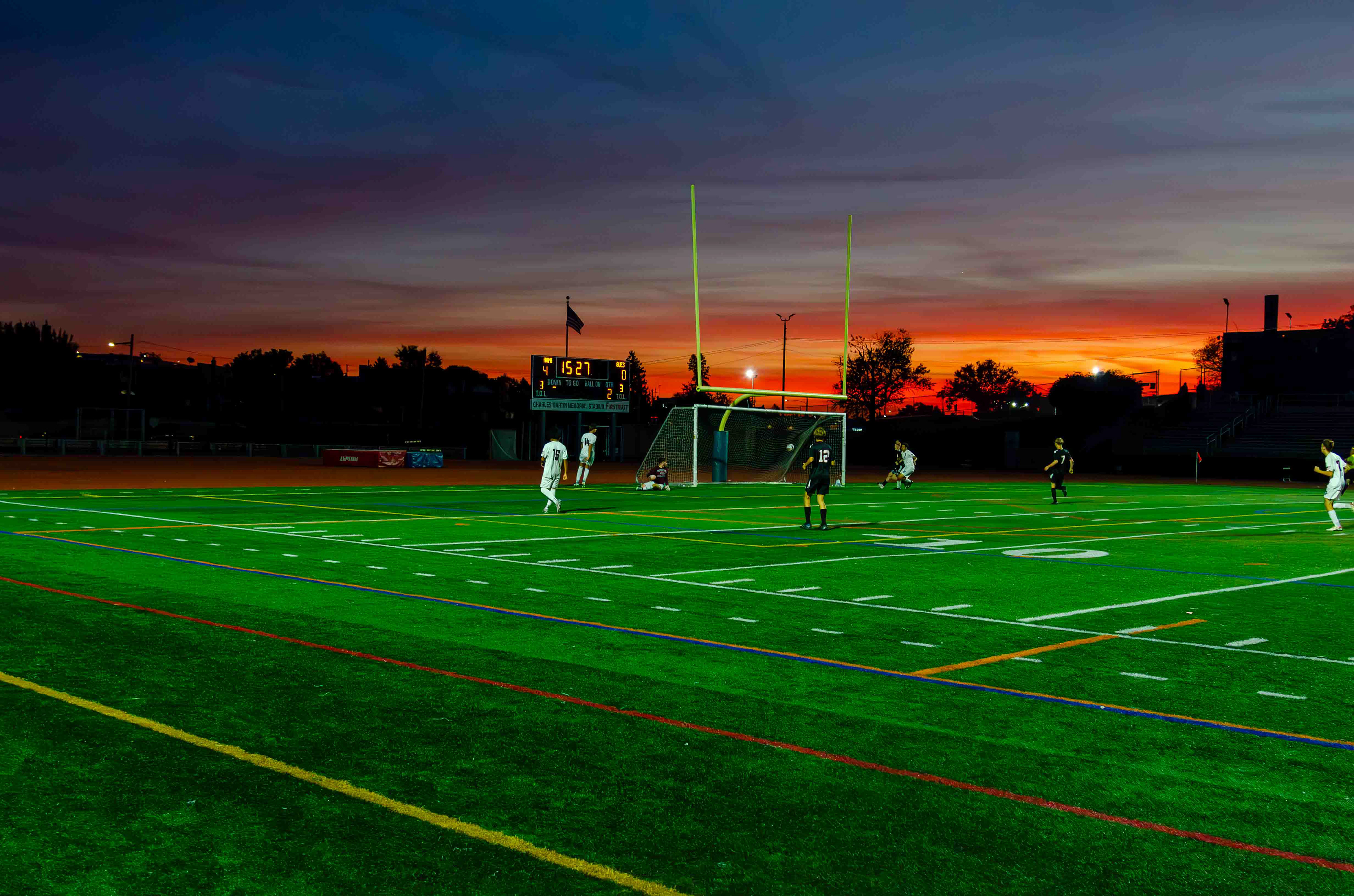 "Cinematic Scale. A wide-angle view of the beautiful game at sunset. Why it’s captivating: * Technically: This is a masterclass in dynamic range. Usually, a bright sunset would turn the foreground into a black silhouette, but here, the turf remains a vibrant, lush green while the sky retains its deep oranges and purples. It shows a sophisticated understanding of exposure compensation.. The leading lines of the field markings and the towering goalposts draw the eye deep into the frame. It evokes the 'Friday Night Lights' feeling—the quiet, epic grandeur that surrounds high school sports."