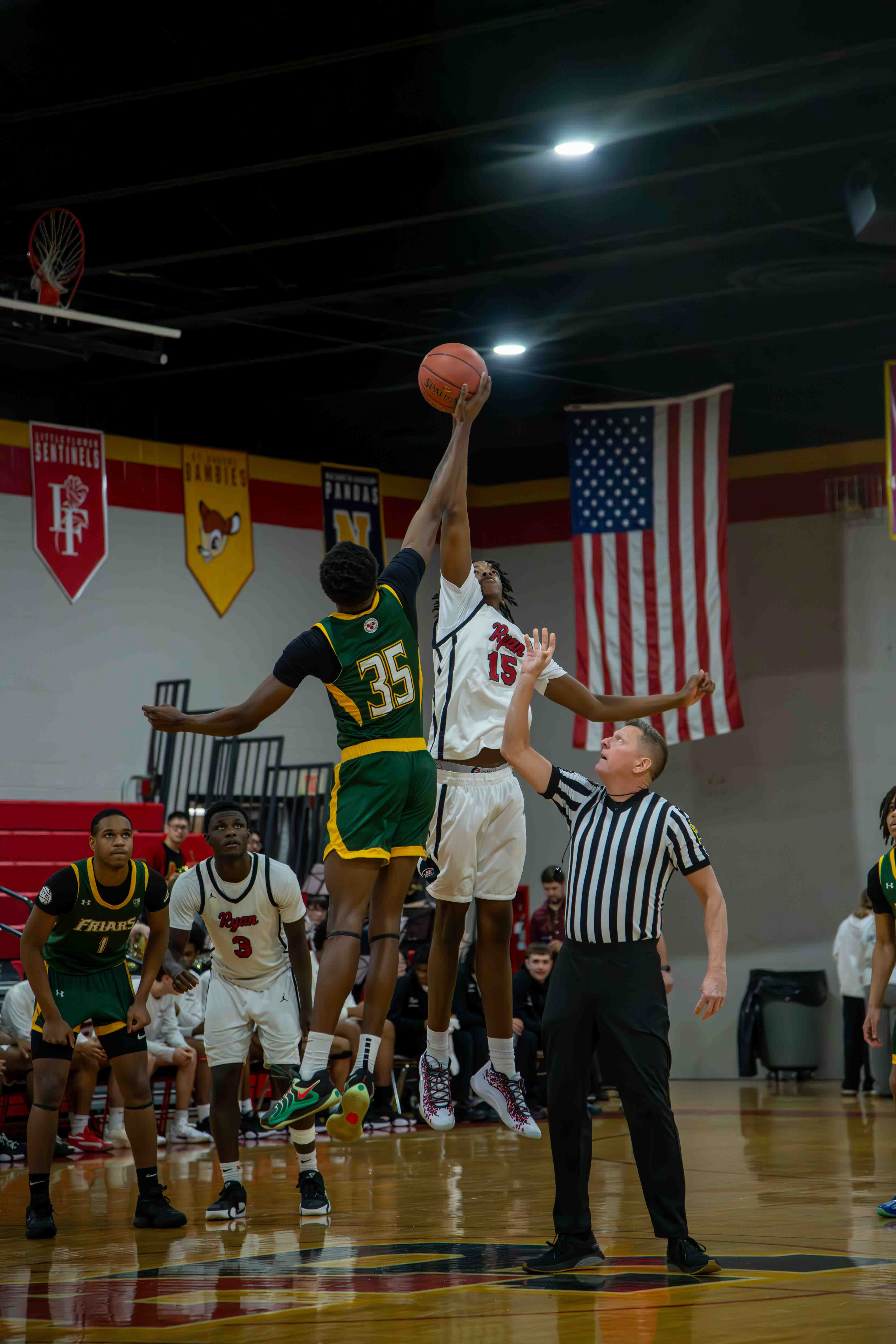 "Timing is everything. This shot captures the exact millisecond of the tip-off, frozen in mid-air. Why it’s captivating: * Technically: Shooting fast action in a high school gym is notoriously difficult due to cycling artificial lights and low-light conditions. This image maintains a tack-sharp focus on the players' hands and the ball, using a high shutter speed and wide aperture to isolate the subjects without losing detail to digital noise.The verticality of the composition creates a sense of monumental scale. The symmetry of the two opposing players reaching for the same goal—framed perfectly against the American flag and the gym banners—tells the story of the entire game in a single frame.