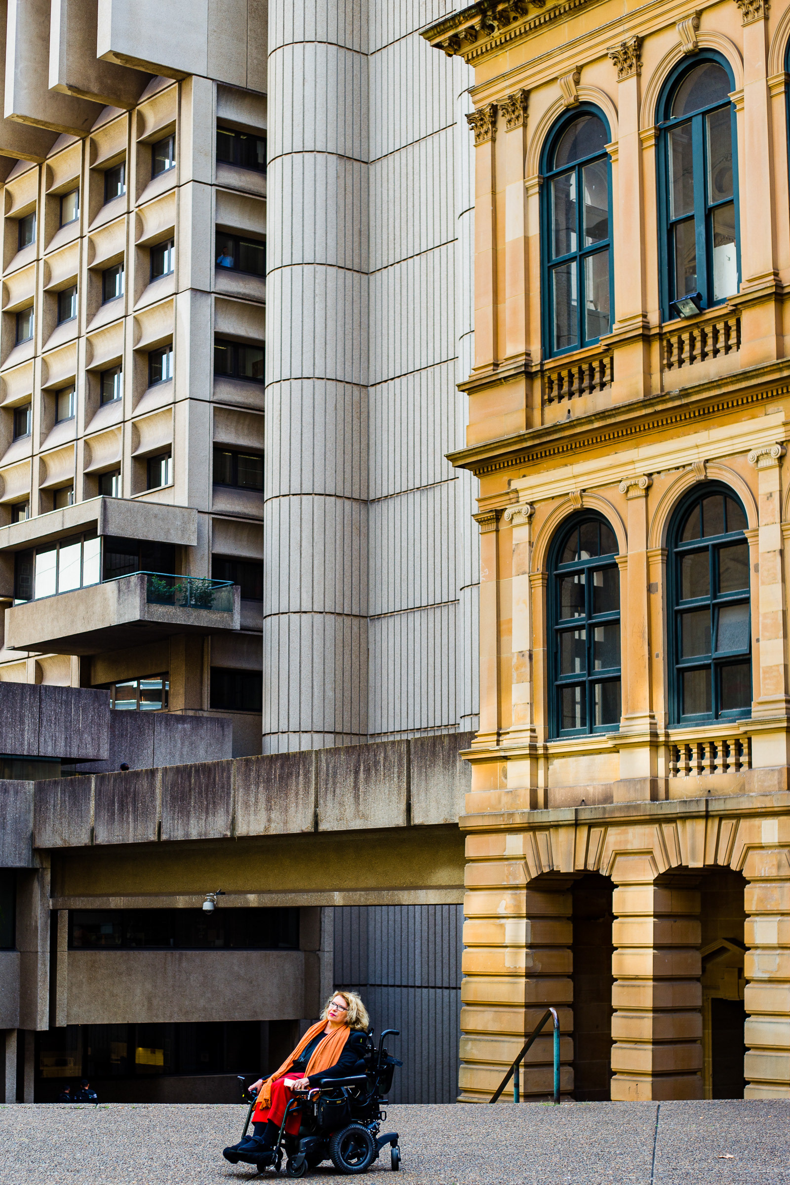 Karen sits alone in her wheelchair wearing black and a large orange scarf, surrounded by architecture in Sydney CBD