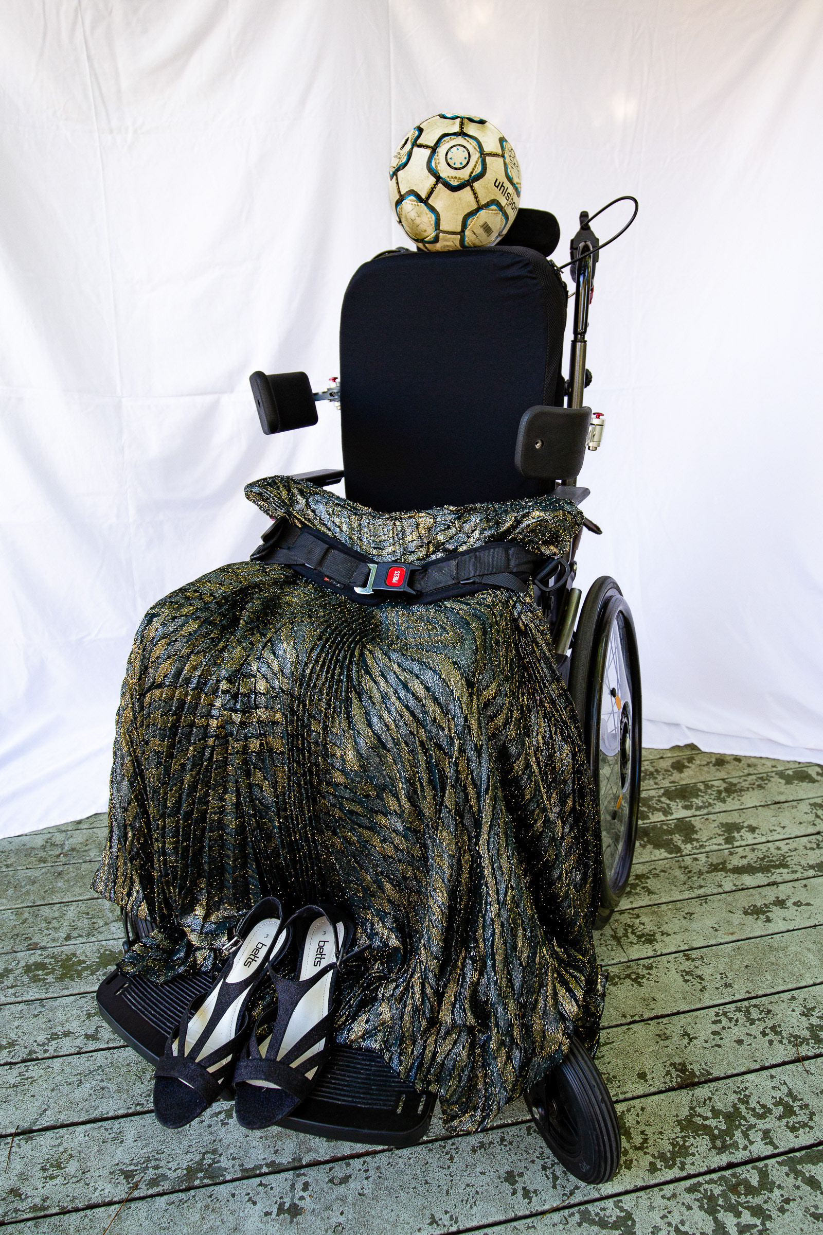 A xheelchair alone, empty with a soccer ball for a head and gold skirt and black heels