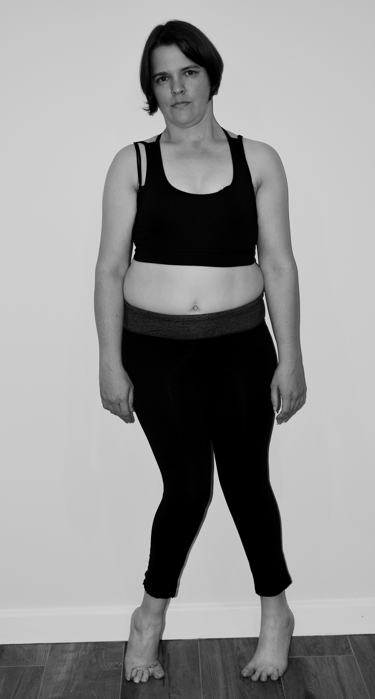 A full frontal photo of Kerry in black and white, against a white wall, wearing black workout gear