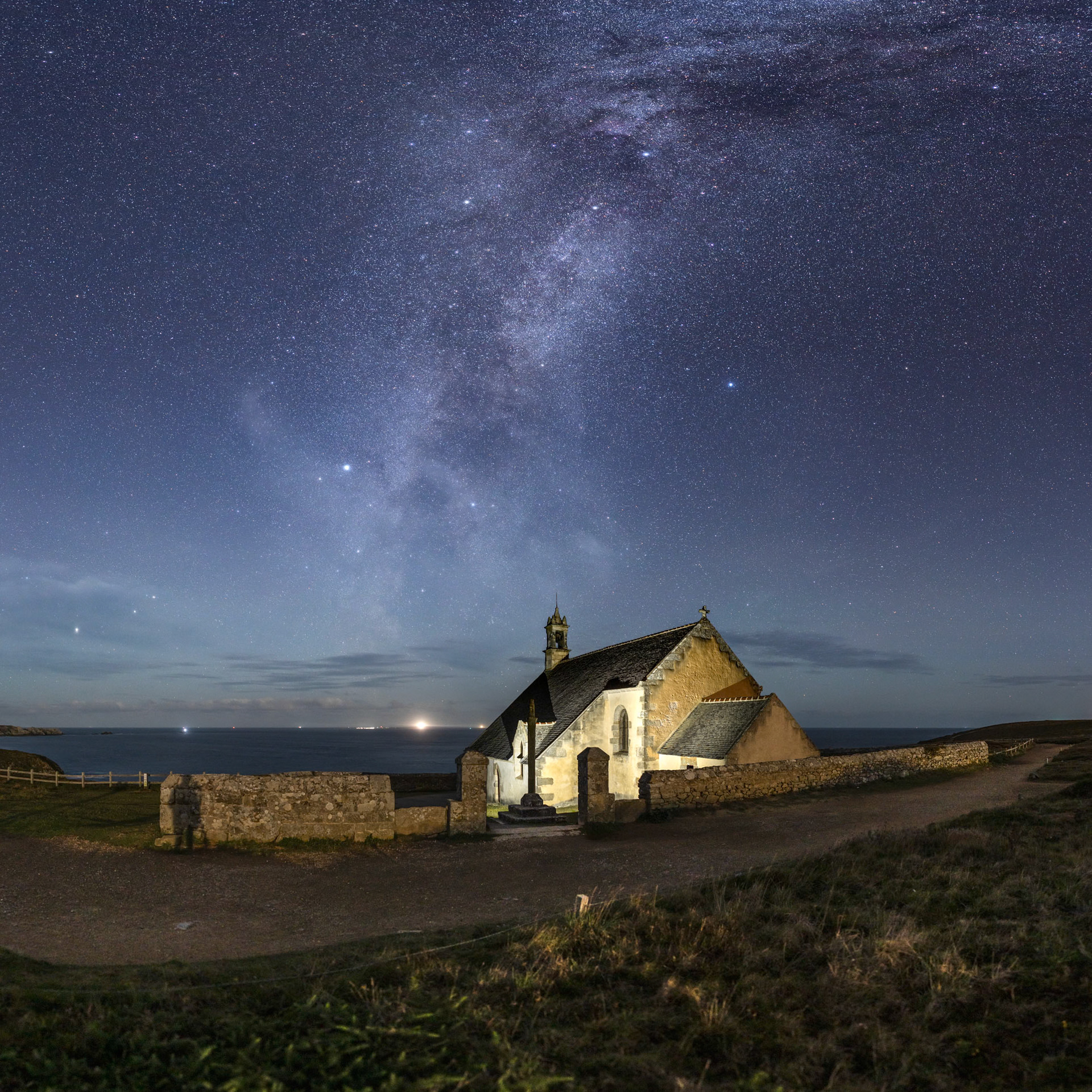 La Voie lactée d'été s'élève à la verticale de l'Île de Sein derrière la chapelle Saint-They érigée à la Pointe du Van. Le flash de mon smartphone installé au pied du calvaire met bien en valeur le volume du bâtiment et dessine chaque ardoise du toit. Mon ombre projetée par le Lune qui se lève dans mon dos est visible tout à gauche du mur d'enceinte.