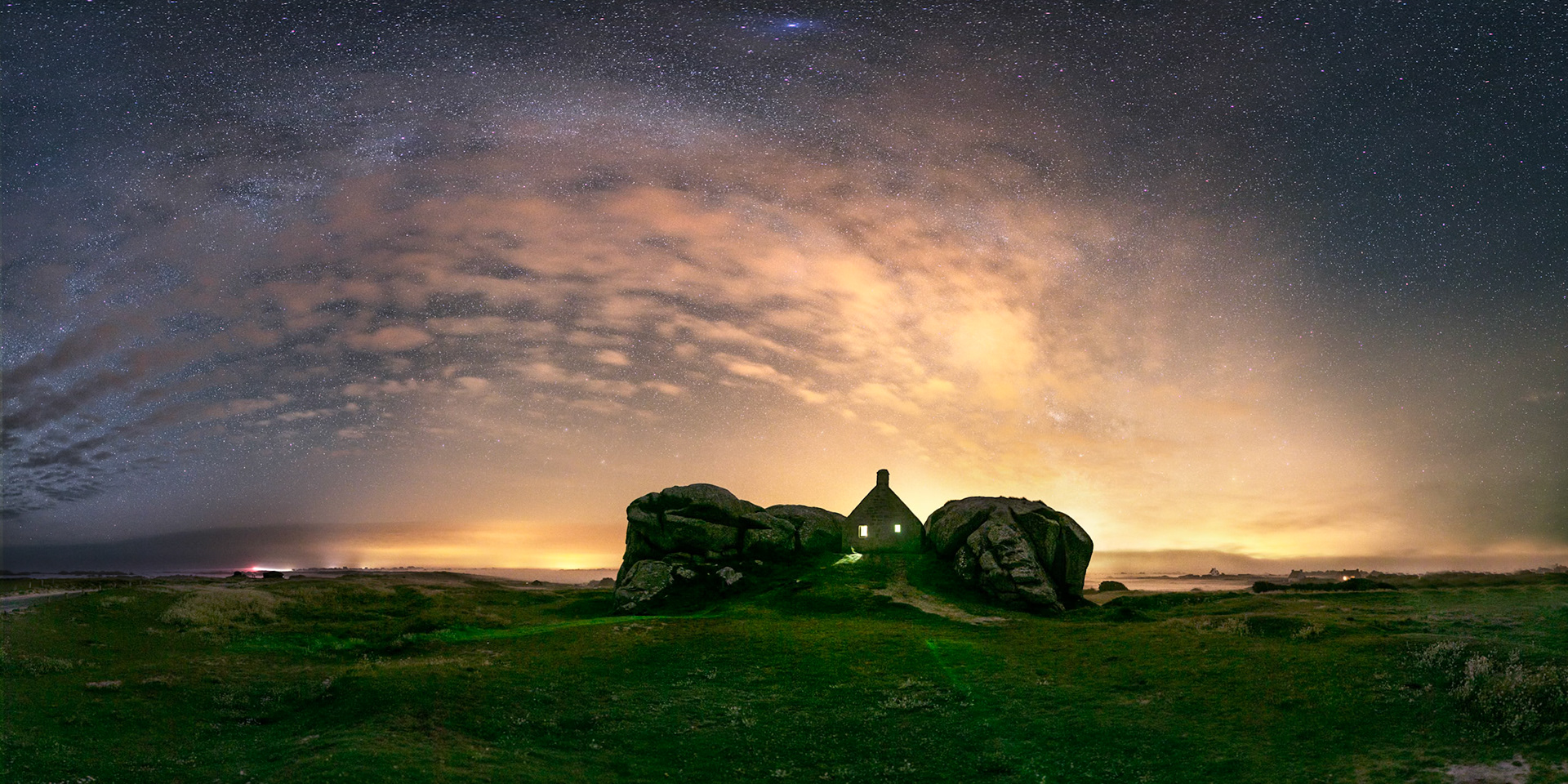Ambiance de légende tout autour de la maison entre les rochers de Menheham (à Kerlouan). Le ciel est empli de nuages qui sont illuminés par les lumières des villes et bourgs environnants (Brest, Lesneven, etc.). Au fond à gauche, la lueur rouge du phare de Pontusval (à Brignogan).