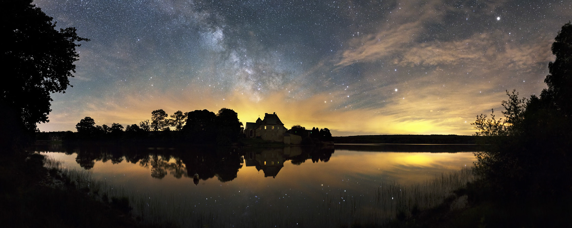 L'Abbaye de Paimpont se reflète dans les eaux calmes de l'Etang de Paimpont. Dans le ciel, une partie de la Voie lactée s'extirpe des cirrus aux teintes orangés (à cause de la pollution lumineuse). A droite de l'Abbaye, le Scorpion et sa brillante Antarès, et plus à droite, Saturne dans la Balance puis Artcurus du Bouvier en haut à droite. EXIF : Canon 6D + Sigma 35mm f/1.4 fermé à 2.0. 10 s à 3200 ISO. Panorama de 23 photos. 3h00.